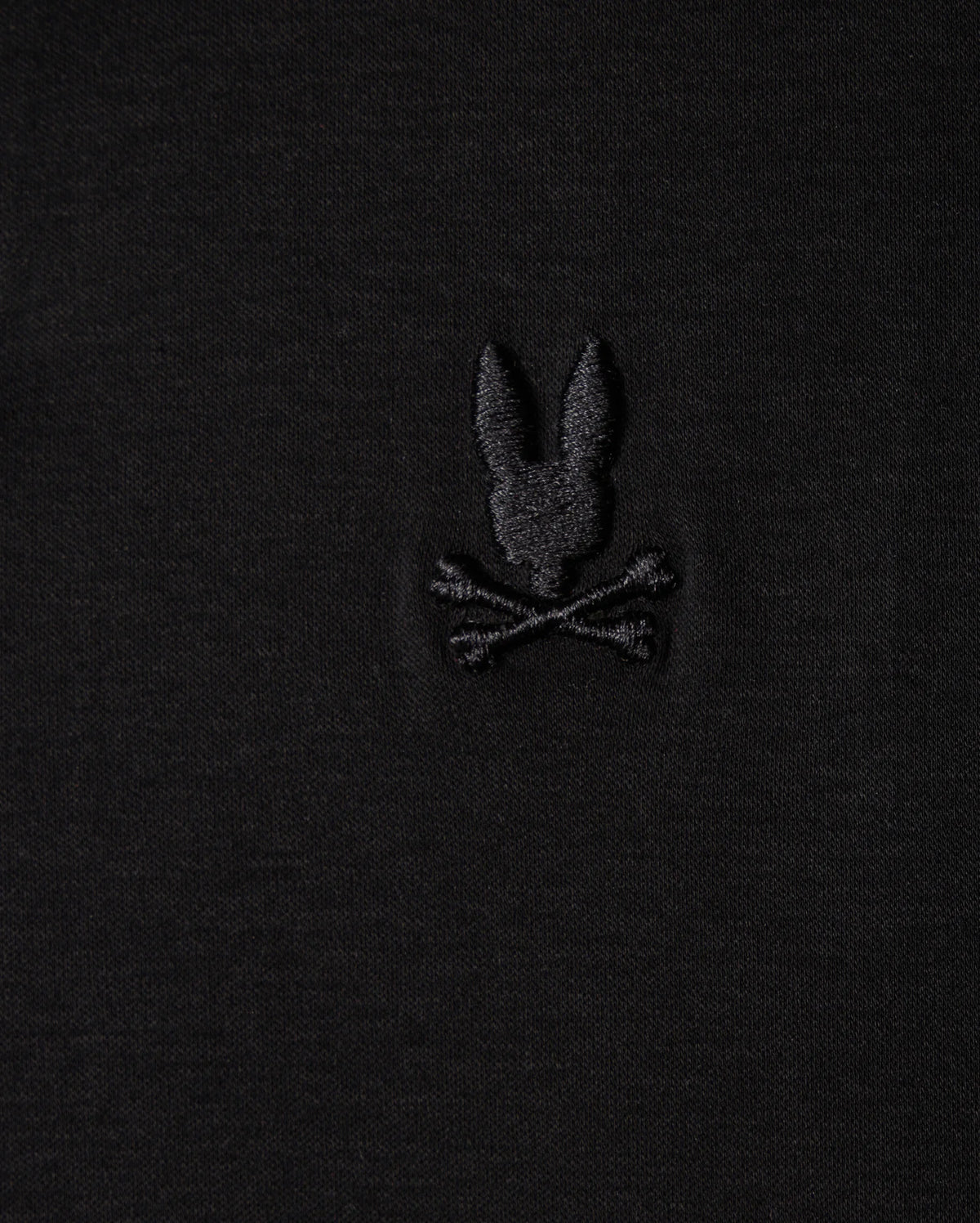 The Psycho Bunny MENS BROADWAY PIMA INTERLOCK JOHNNY COLLAR POLO - B6K891D200 is a black fabric polo made from durable lustre Mercerized Pima cotton. It features a subtle embroidered design of a stylized rabbit head above two crossed bones, evoking the classic skull and crossbones motif.