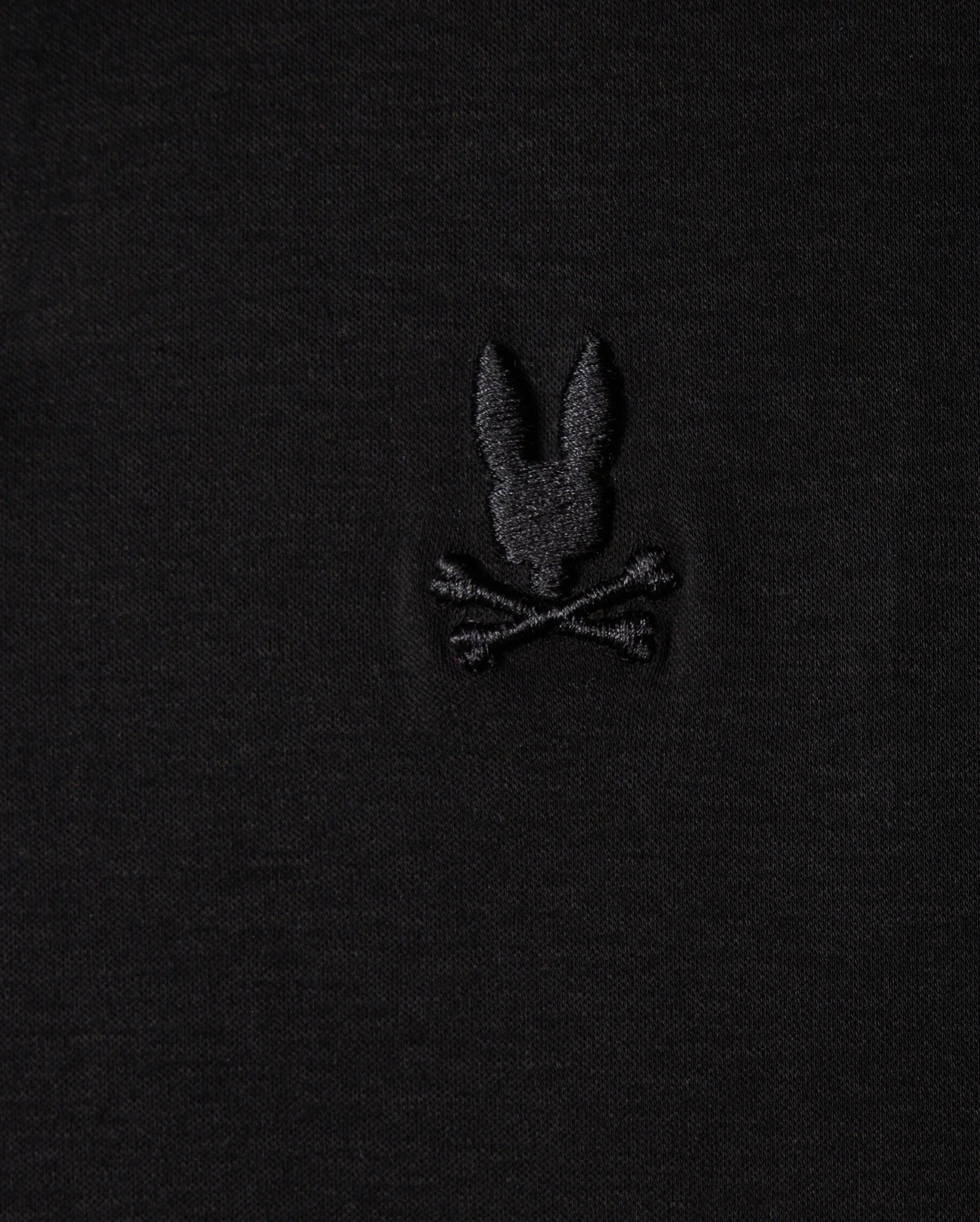 The Psycho Bunny MENS BROADWAY PIMA INTERLOCK JOHNNY COLLAR POLO - B6K891D200 is a black fabric polo made from durable lustre Mercerized Pima cotton. It features a subtle embroidered design of a stylized rabbit head above two crossed bones, evoking the classic skull and crossbones motif.