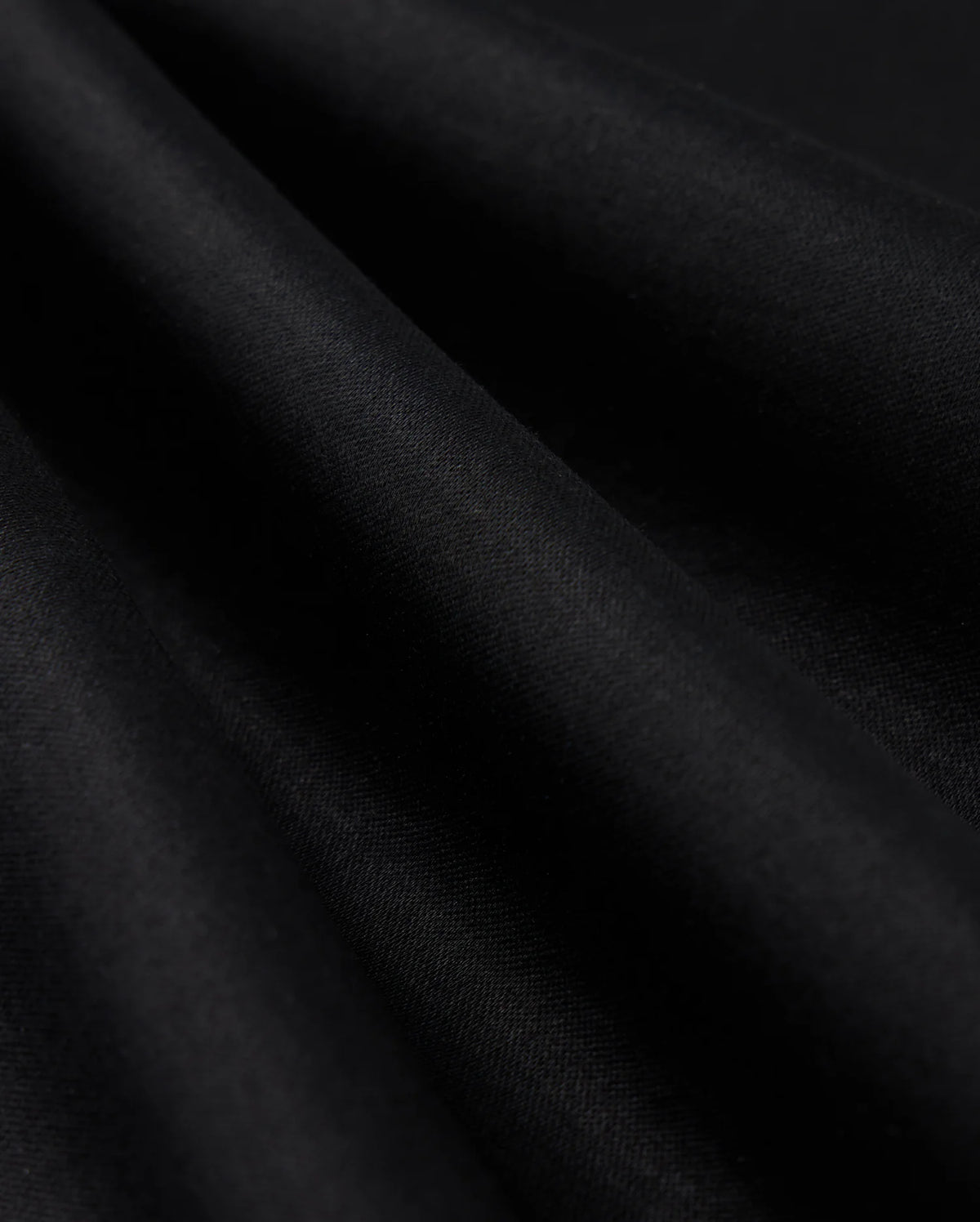 Close-up of the smooth, dark fabric of the Psycho Bunny MENS BROADWAY PIMA INTERLOCK JOHNNY COLLAR POLO - B6K891D200, with gentle folds creating subtle shadows and highlights. The Mercerized Pima cotton demonstrates a durable lustre, appearing soft and draped, showcasing its texture and depth against a dark background.
