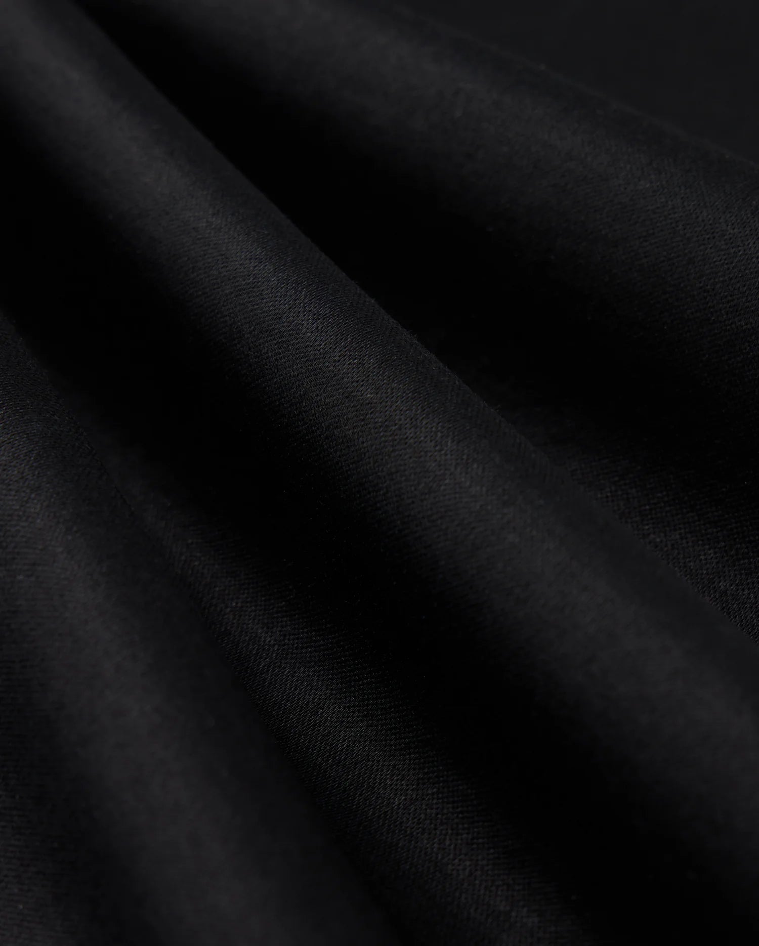 Close-up of the smooth, dark fabric of the Psycho Bunny MENS BROADWAY PIMA INTERLOCK JOHNNY COLLAR POLO - B6K891D200, with gentle folds creating subtle shadows and highlights. The Mercerized Pima cotton demonstrates a durable lustre, appearing soft and draped, showcasing its texture and depth against a dark background.