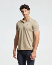 A man wearing a Psycho Bunny MENS BROADWAY PIMA INTERLOCK JOHNNY COLLAR POLO - B6K891D200 in beige stands against a plain light background. His hair is curly, and he looks directly at the camera, pairing his outfit with dark pants.