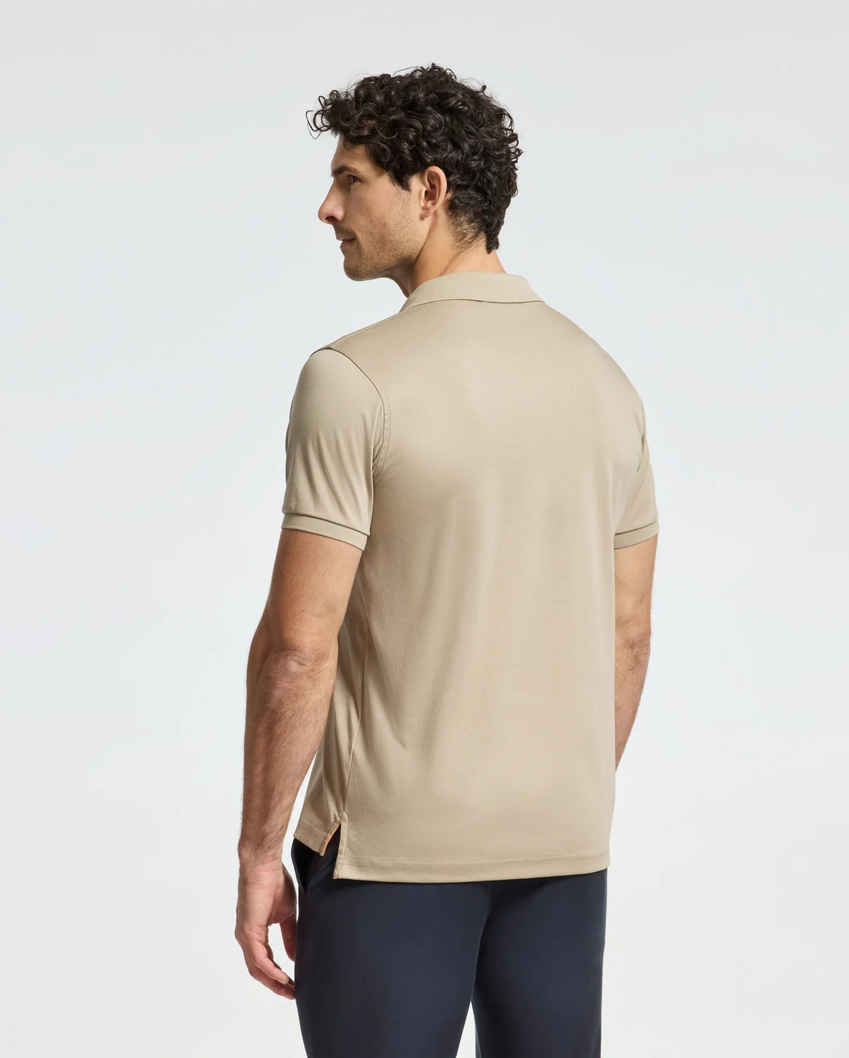 A man with curly hair, in a MENS BROADWAY PIMA INTERLOCK JOHNNY COLLAR POLO by Psycho Bunny and dark pants, stands facing away from the camera against a light grey background.