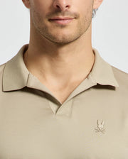 A man wearing a Psycho Bunny MENS BROADWAY PIMA INTERLOCK JOHNNY COLLAR POLO in beige, featuring a small embroidered logo of a rabbit and crossbones on the chest. The focus is on the upper torso and the shirt.