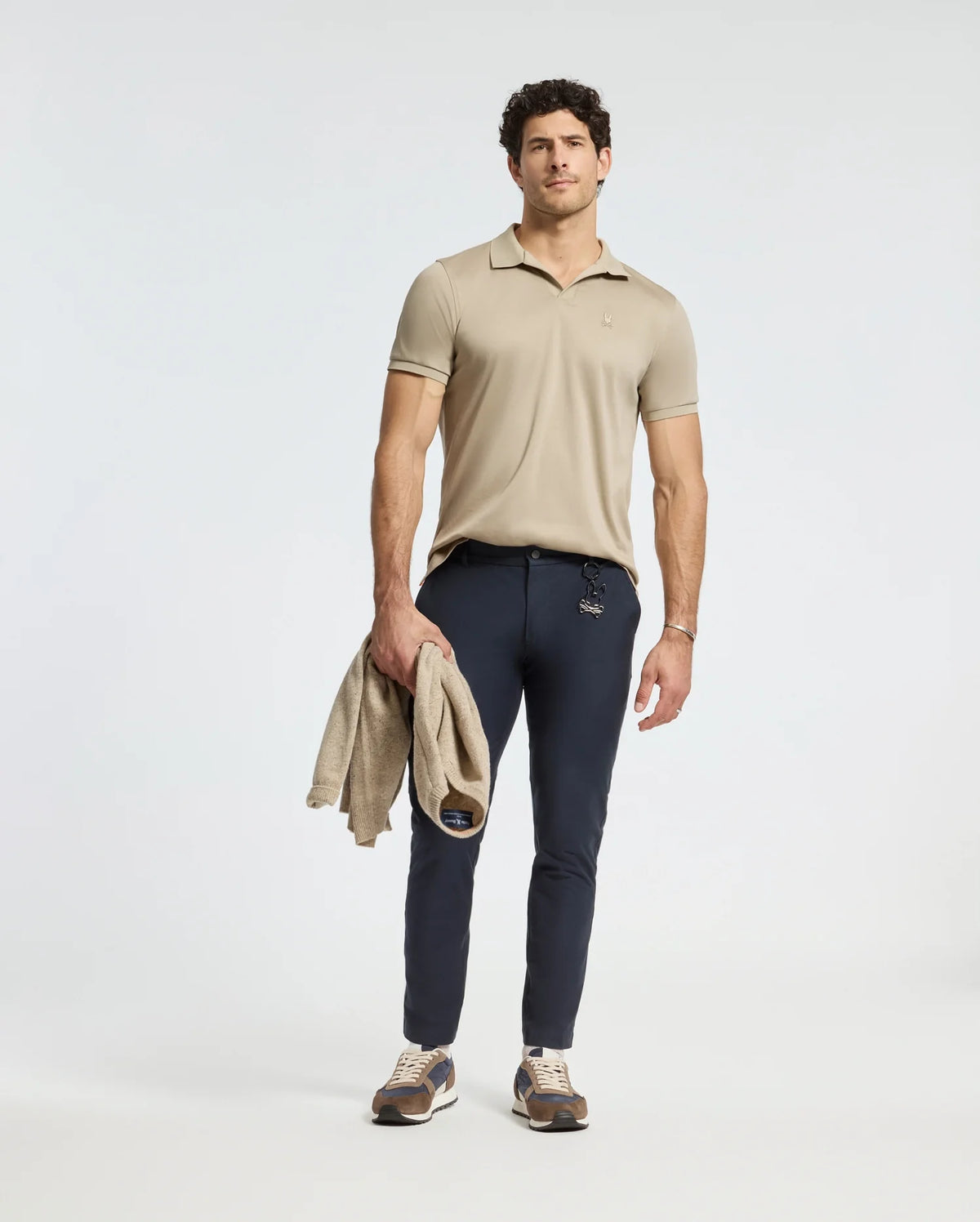 A man stands wearing the Psycho Bunny Mens Broadway Pima Interlock Johnny Collar Polo in beige, paired with navy pants and sneakers. He holds a matching beige sweater in his left hand against a plain, light-colored background.