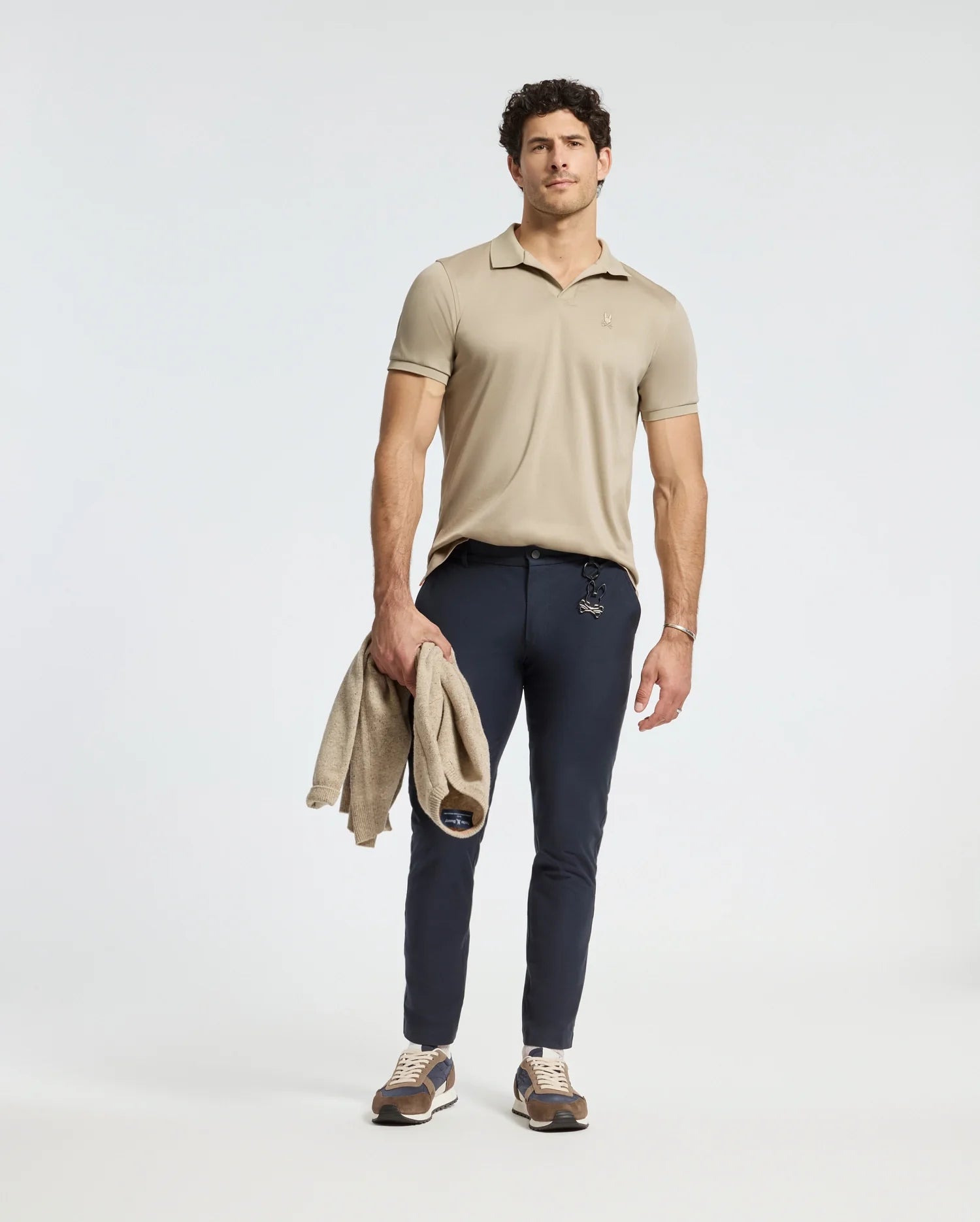 A man stands wearing the Psycho Bunny Mens Broadway Pima Interlock Johnny Collar Polo in beige, paired with navy pants and sneakers. He holds a matching beige sweater in his left hand against a plain, light-colored background.
