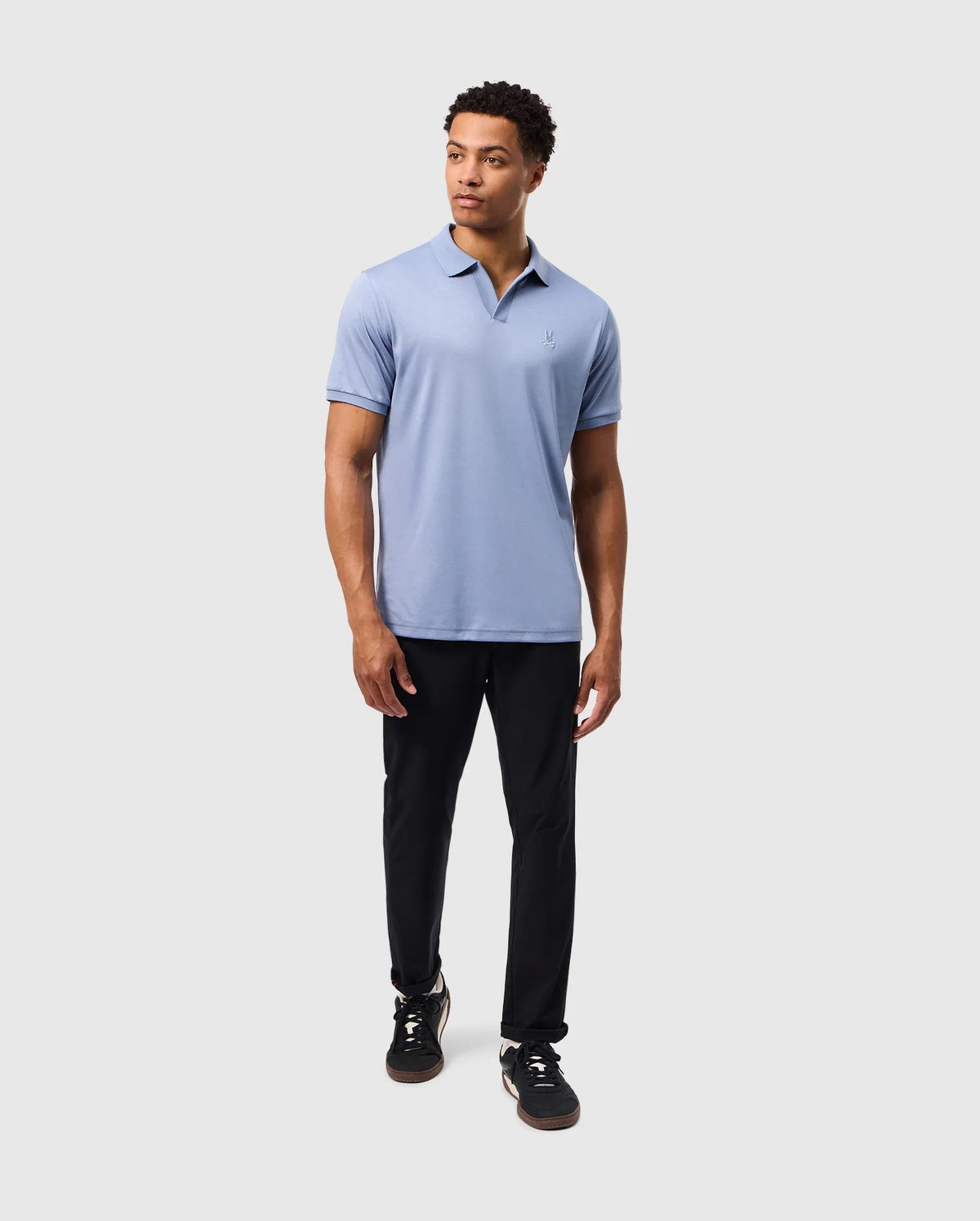 A person in a light blue Psycho Bunny Broadway Mercerized Pima Polo (B6K891D200) paired with black pants stands against a gray background. With short hair and black sneakers, they gaze to the side, their attire reflecting a hint of durable lustre.