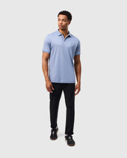 A person in a light blue Psycho Bunny Broadway Mercerized Pima Polo (B6K891D200) paired with black pants stands against a gray background. With short hair and black sneakers, they gaze to the side, their attire reflecting a hint of durable lustre.