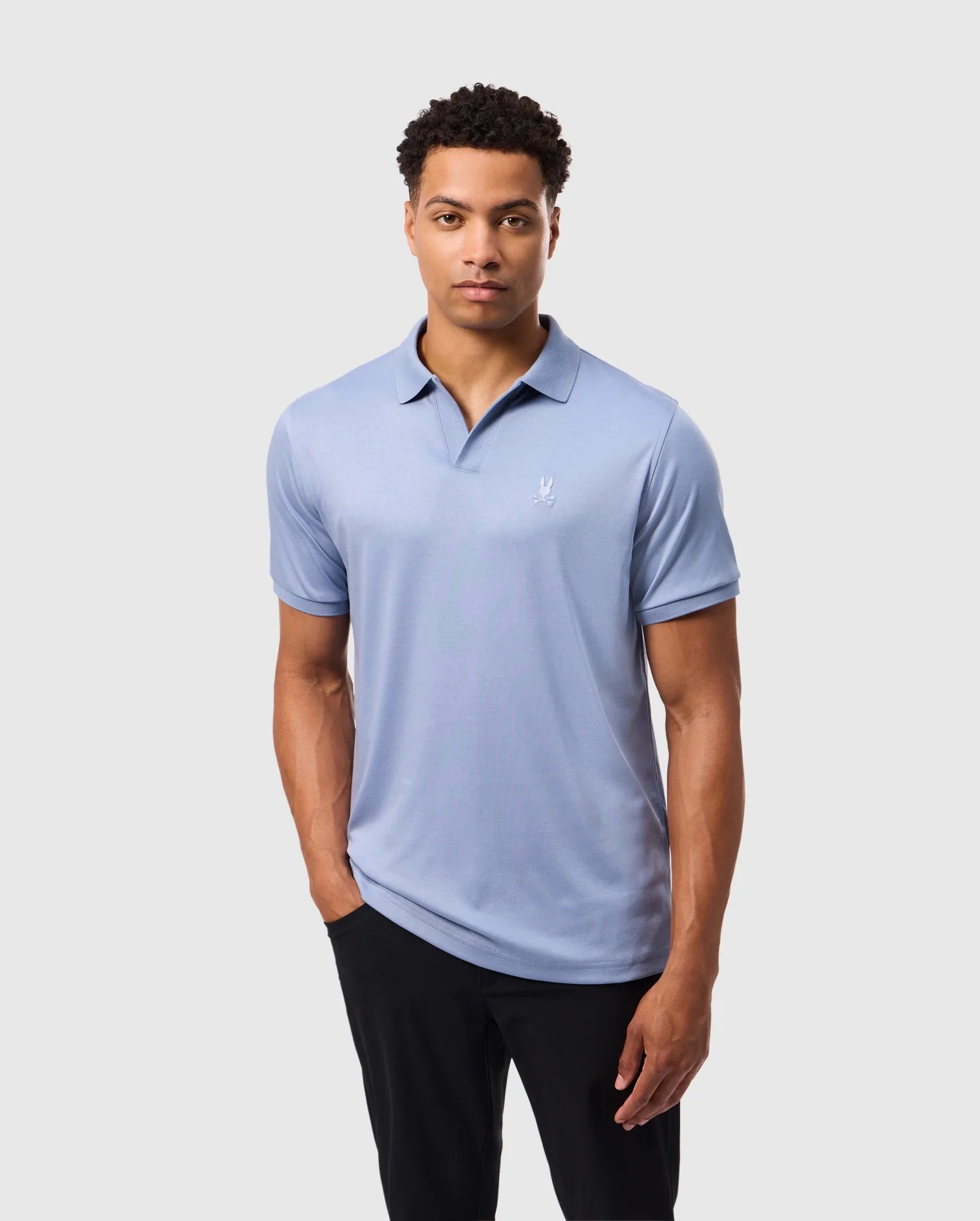 A man in a Psycho Bunny MENS BROADWAY MERCERIZED PIMA POLO SHIRT (B6K891D200), light blue with a small white logo, stands against a plain white background. Made from durable, lustrous mercerized Pima cotton, he is paired with black pants and has short curly hair and a neutral expression.