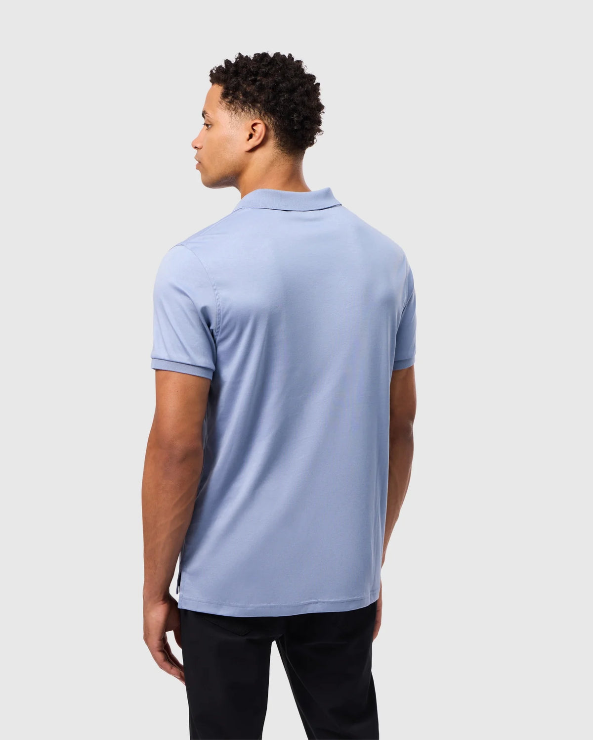 A man with short curly hair wears a Psycho Bunny Men's Broadway Mercerized Pima Polo Shirt (B6K891D200) in light blue, paired with black pants. He stands with his back turned, looking right against a plain gray background.