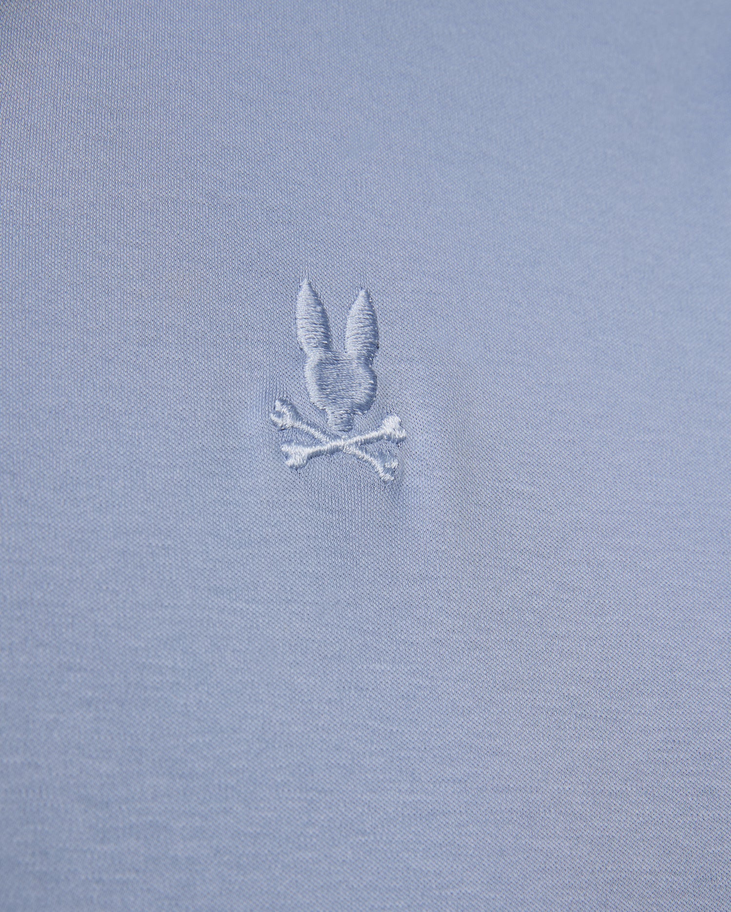 The MENS BROADWAY MERCERIZED PIMA POLO SHIRT by Psycho Bunny showcases a close-up of durable gray luster fabric with an embroidered design featuring a bunny head above two crossed bones.
