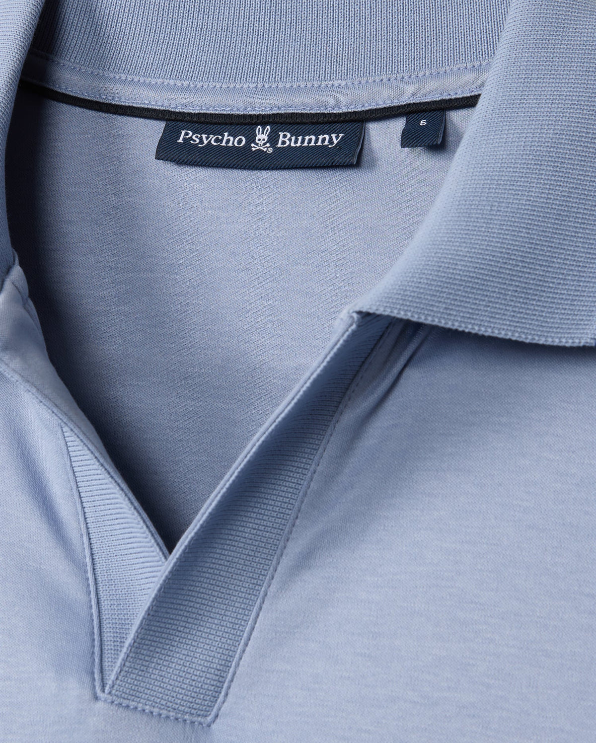 Close-up of the MENS BROADWAY MERCERIZED PIMA POLO SHIRT - B6K891D200 in light blue, featuring the "Psycho Bunny" black label at the collar with a bunny emblem. The durable lustre of the mercerized Pima cotton enhances its texture, highlighting stitching details for elegance and resilience.
