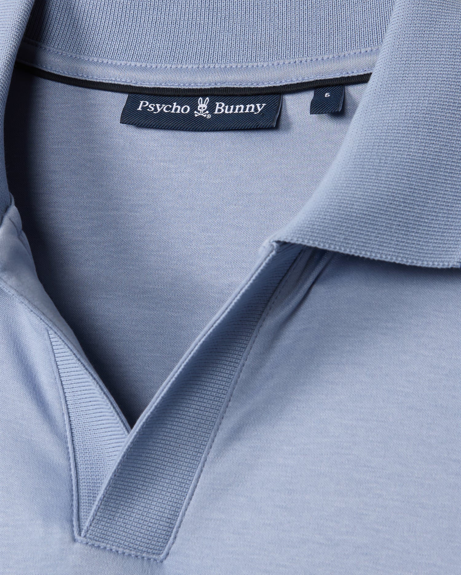 Close-up of the MENS BROADWAY MERCERIZED PIMA POLO SHIRT - B6K891D200 in light blue, featuring the "Psycho Bunny" black label at the collar with a bunny emblem. The durable lustre of the mercerized Pima cotton enhances its texture, highlighting stitching details for elegance and resilience.