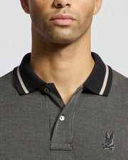 A close-up of a person wearing the Psycho Bunny "MENS WARSAW JACQUARD PIQUE POLO - B6K901E200" crafted from Peruvian Pima cotton, featuring a black and grey jacquard-knit design with a subtle checkered pattern. The polo shirt is accented with a black collar adorned with white stripes, and it has the brand's distinctive small embroidered logo of a rabbit with crossed bones. The person's face is partially visible in the image.