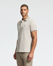 A man dressed in a light gray Psycho Bunny Men's Warsaw Jacquard Pique Polo, made from luxurious Peruvian Pima cotton and featuring an embroidered logo on the chest, stands against a plain white background. He has short dark hair and light stubble, with his hands by his sides, looking confidently at the camera.