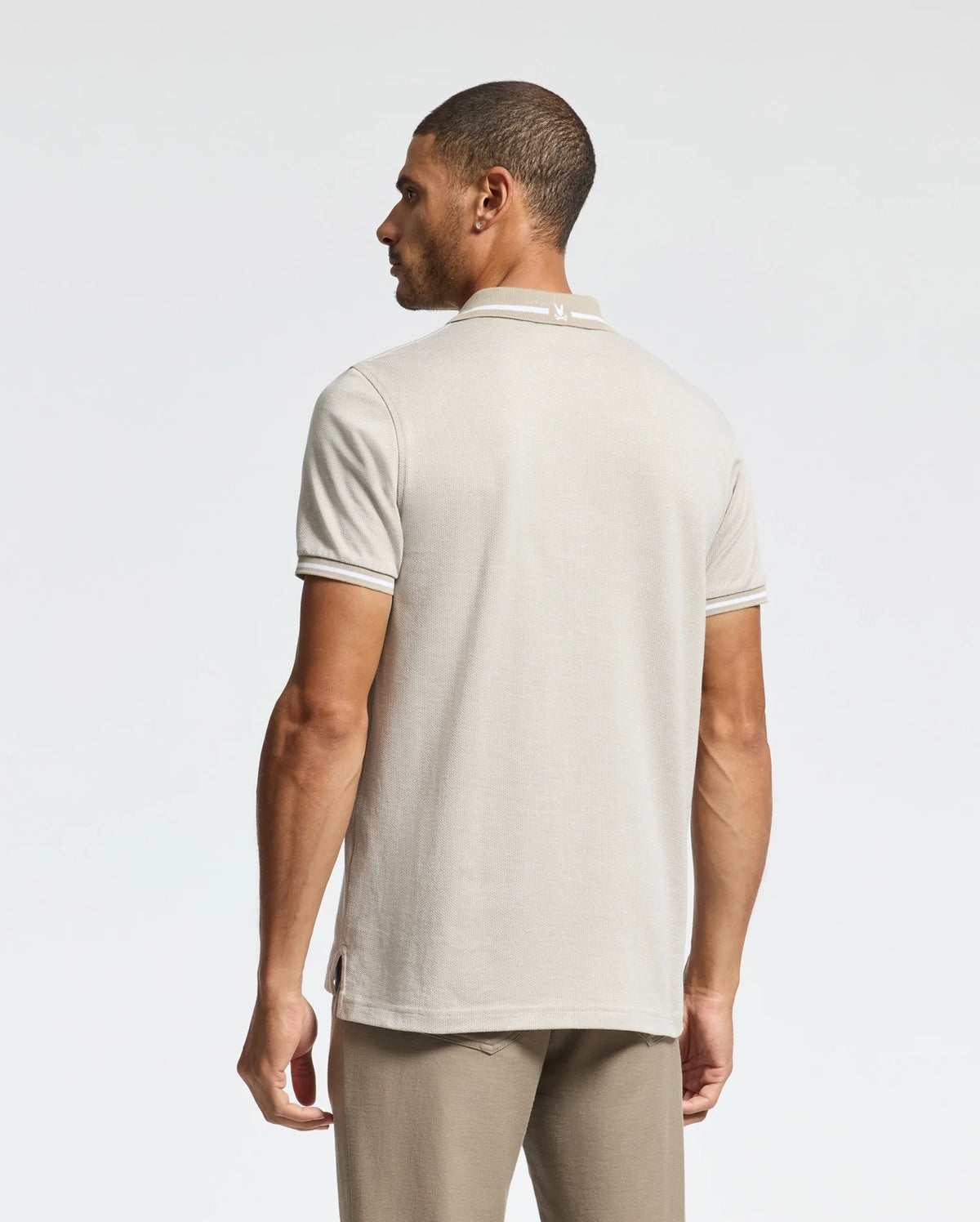 A man is seen from behind wearing a Psycho Bunny MENS WARSAW JACQUARD PIQUE POLO in light beige, crafted from luxurious Peruvian Pima cotton. The polo showcases a subtle collar design and pairs seamlessly with his pants against the plain white backdrop.