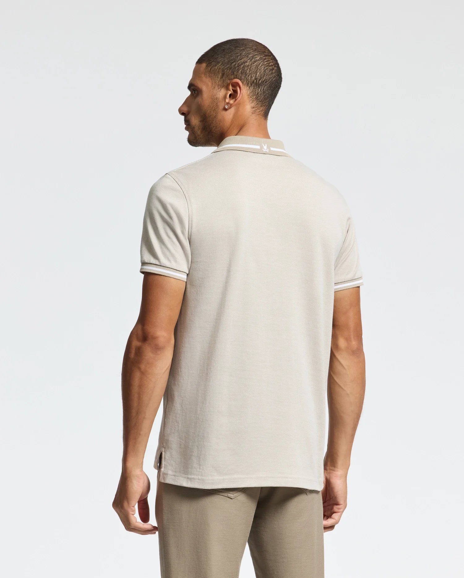 A man is seen from behind wearing a Psycho Bunny MENS WARSAW JACQUARD PIQUE POLO in light beige, crafted from luxurious Peruvian Pima cotton. The polo showcases a subtle collar design and pairs seamlessly with his pants against the plain white backdrop.