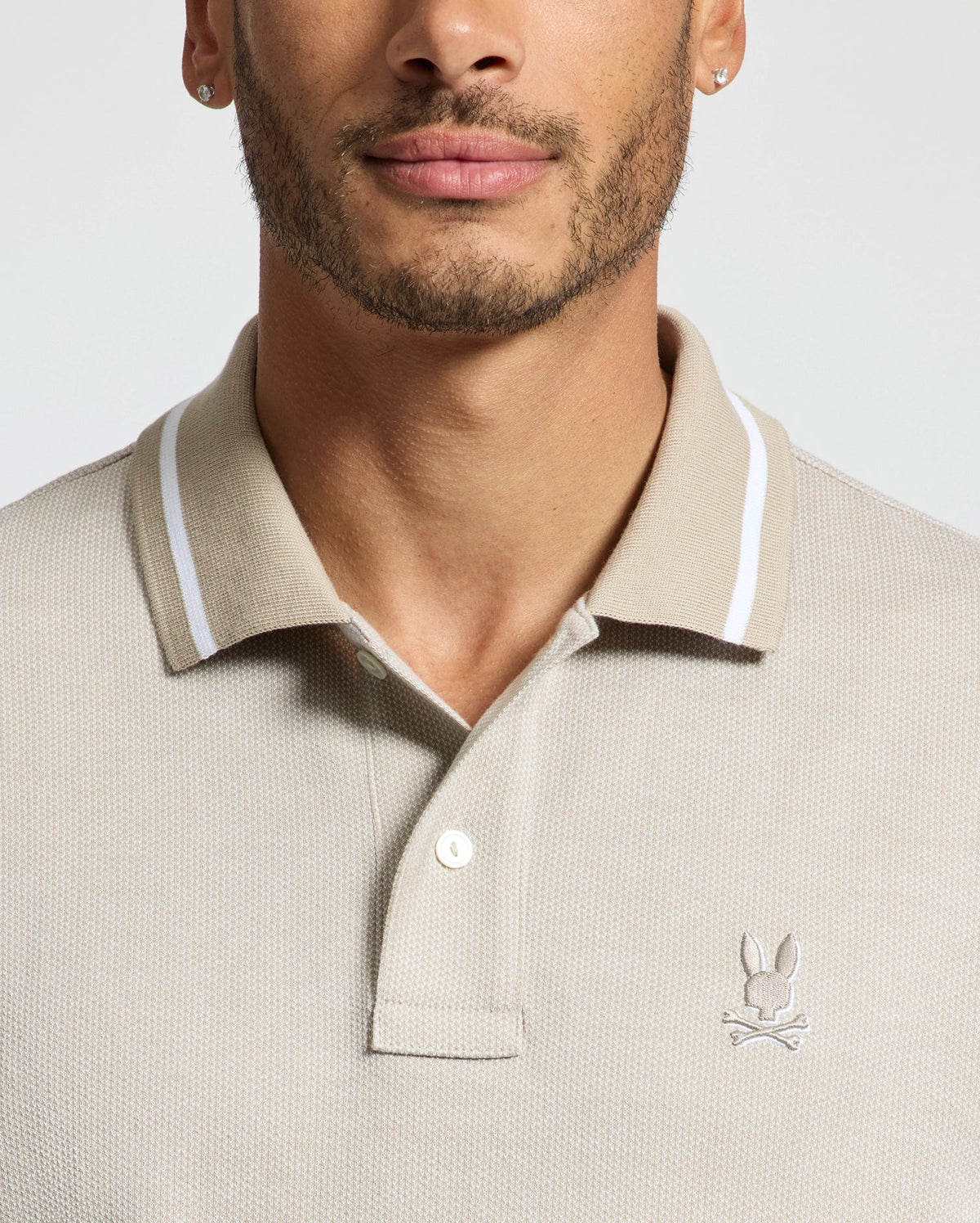 A man is wearing the Psycho Bunny Men's Warsaw Jacquard Pique Polo, made from soft Peruvian Pima cotton. It features a white collar trim and the iconic bunny logo on the chest. He also has earrings and a trimmed beard, with the photo cropped below his nose and above his elbows against a plain background.