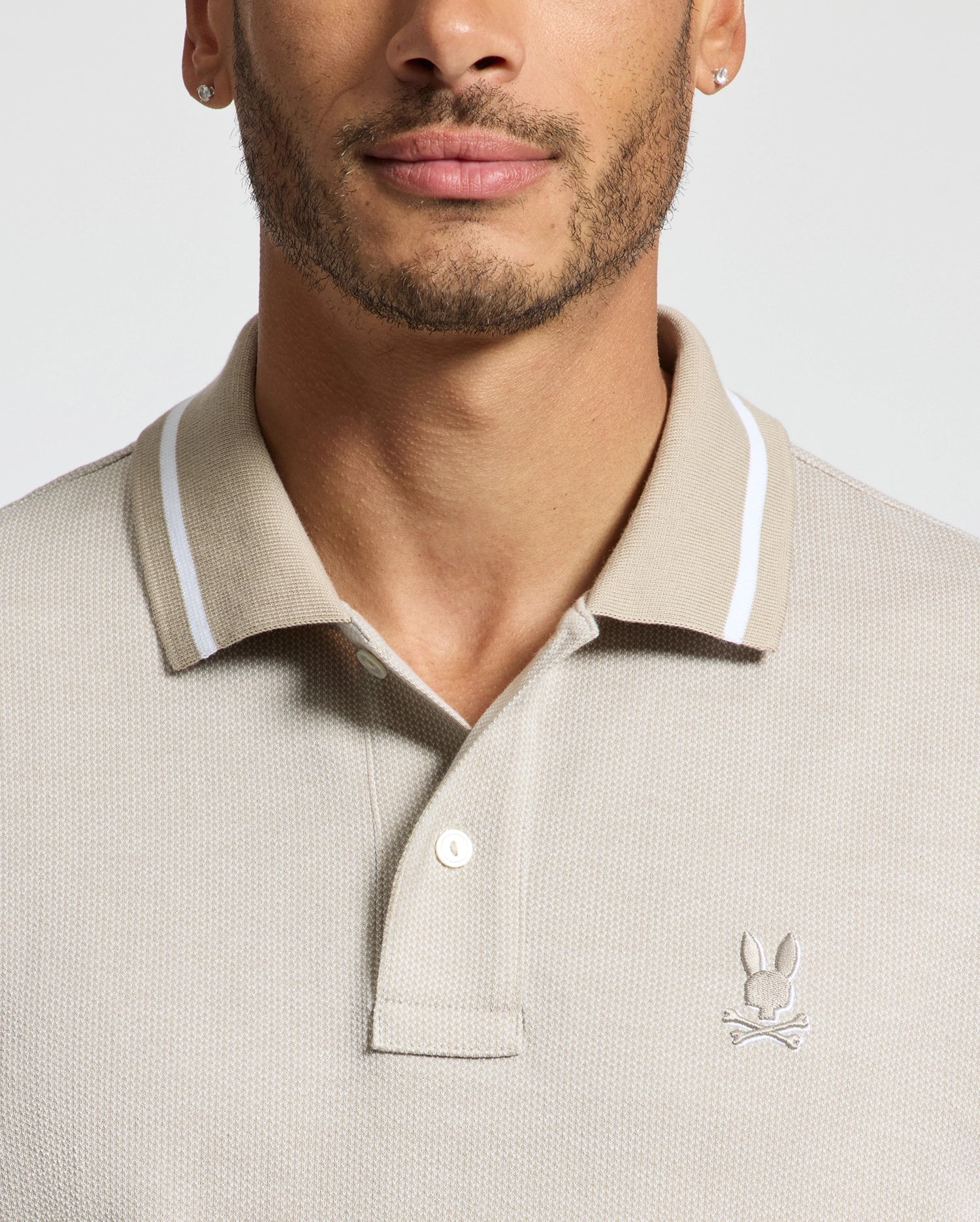 A man is wearing the Psycho Bunny Men's Warsaw Jacquard Pique Polo, made from soft Peruvian Pima cotton. It features a white collar trim and the iconic bunny logo on the chest. He also has earrings and a trimmed beard, with the photo cropped below his nose and above his elbows against a plain background.