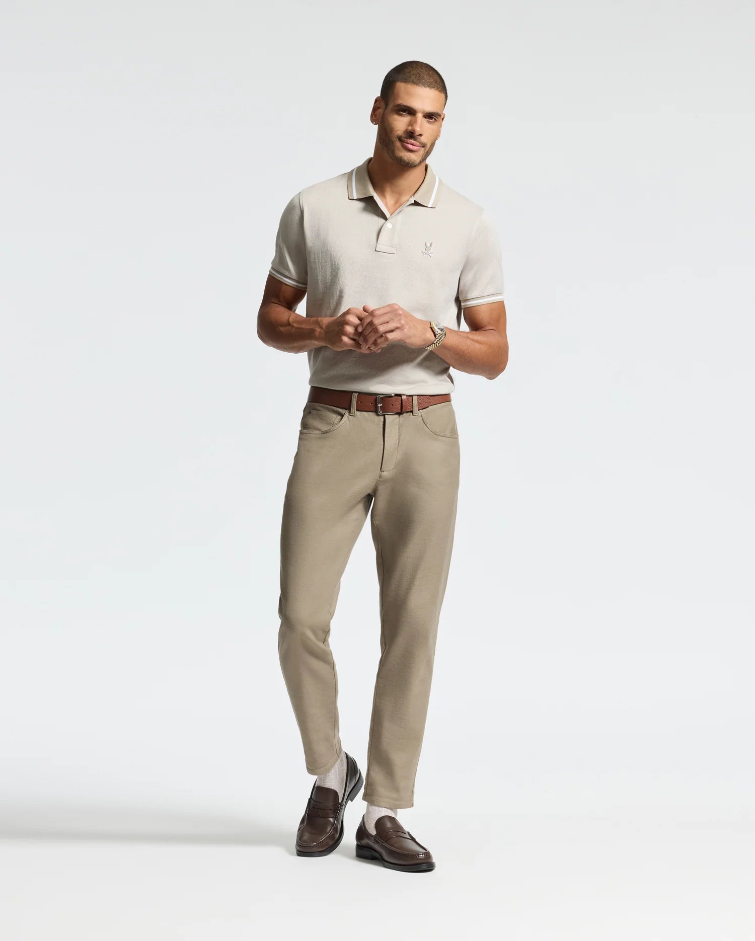 A man stands against a plain backdrop wearing a light beige Psycho Bunny MENS WARSAW JACQUARD PIQUE POLO - B6K901E200, crafted from Peruvian Pima cotton. He pairs it with khaki pants and brown loafers that match his belt. Casually posing, he places one hand on his belt while the other hangs freely by his side.