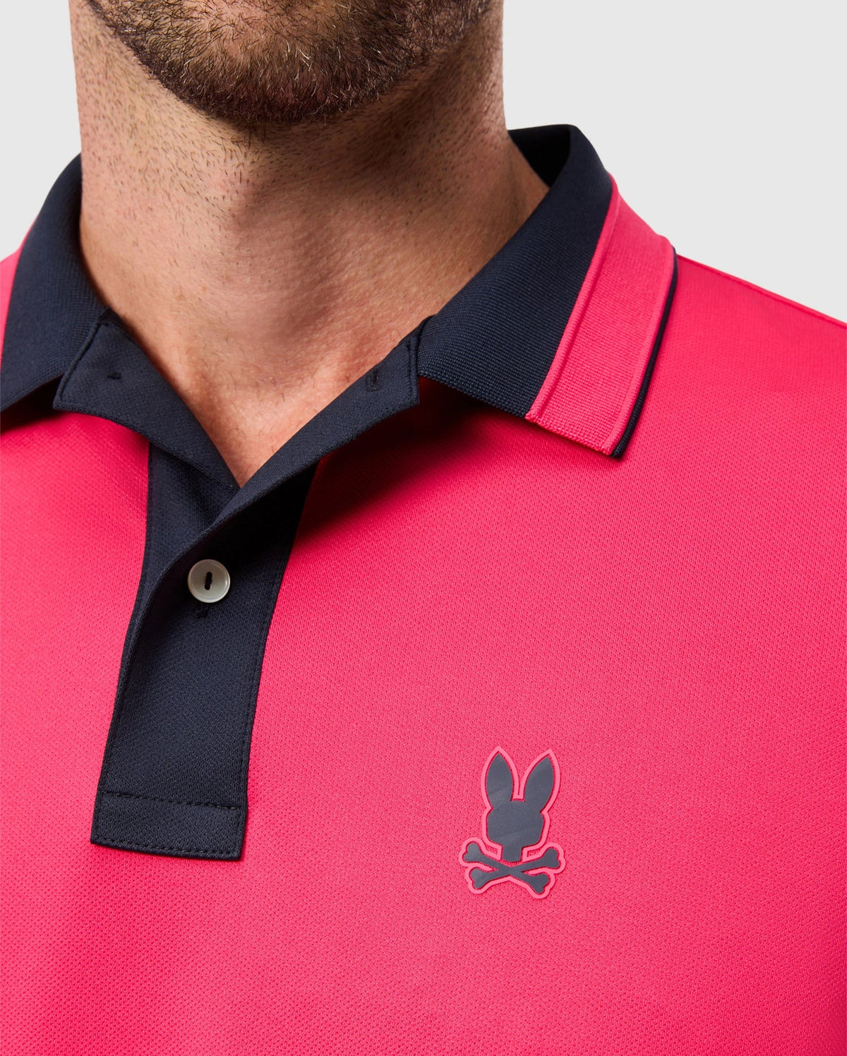 A person wearing the MENS BELLEVUE SPORT POLO SHIRT - B6K905D200 by Psycho Bunny, featuring a versatile casual design in red with a black collar and button placket. The shirt displays the iconic logo of a bunny head above crossed bones on the chest and is crafted as an athletic knit polo for men, making it perfect for any occasion against the plain background.
