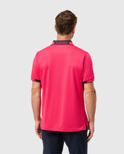 A man stands with his back to the camera, showcasing a stylish MENS BELLEVUE SPORT POLO SHIRT by Psycho Bunny. The shirt features a vibrant bright pink color with black trim and text detailing on the collar. He pairs it with dark trousers, all set against a plain light gray backdrop that emphasizes the effortless elegance of his ensemble.
