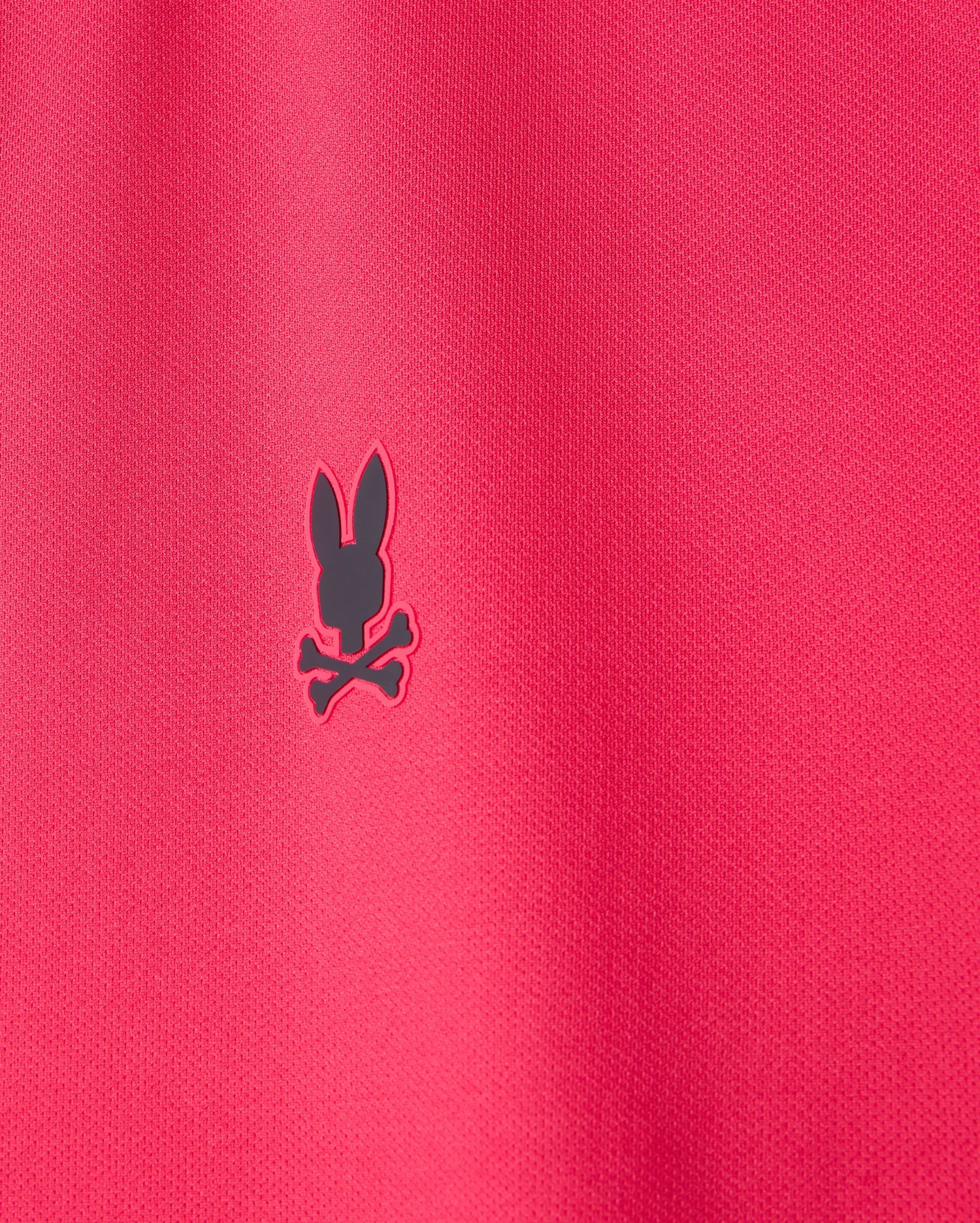 Close-up of a red fabric, typical of the breathable MENS BELLEVUE SPORT POLO SHIRT - B6K905D200 by Psycho Bunny, with a small black logo showcasing a rabbit head and crossbones.