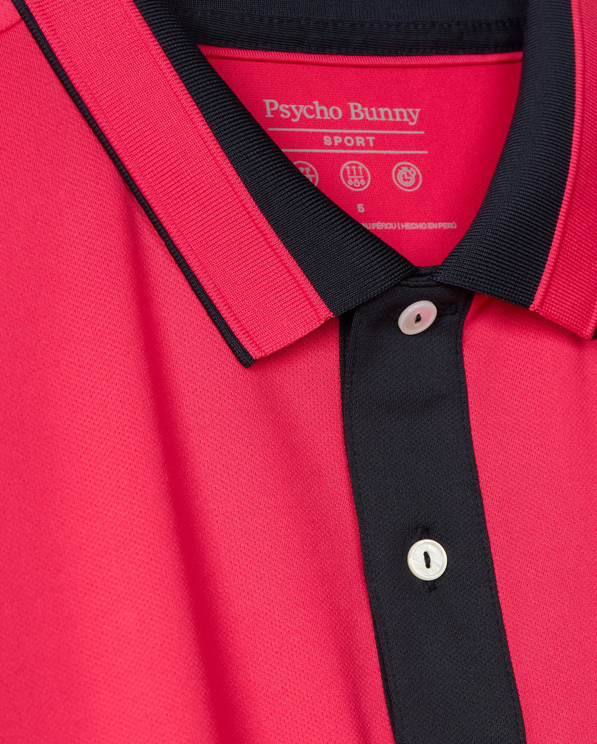 A close-up of the versatile casual pink Bellevue Sport Polo Shirt by Psycho Bunny, showcasing its black trim on the collar and button placket with visible white buttons. The breathable performance polo features a tag that indicates the brand name "Psycho Bunny.