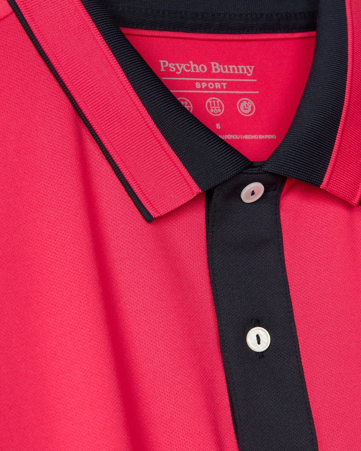A close-up of the versatile casual pink Bellevue Sport Polo Shirt by Psycho Bunny, showcasing its black trim on the collar and button placket with visible white buttons. The breathable performance polo features a tag that indicates the brand name "Psycho Bunny.