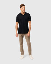 A man stands against a plain background, wearing the Psycho Bunny MENS JAMES PREMIUM PIQUE POLO SHIRT - B6K945D200 in black, paired with beige pants and white sneakers. His short hair is neatly styled as he gazes slightly to the side.