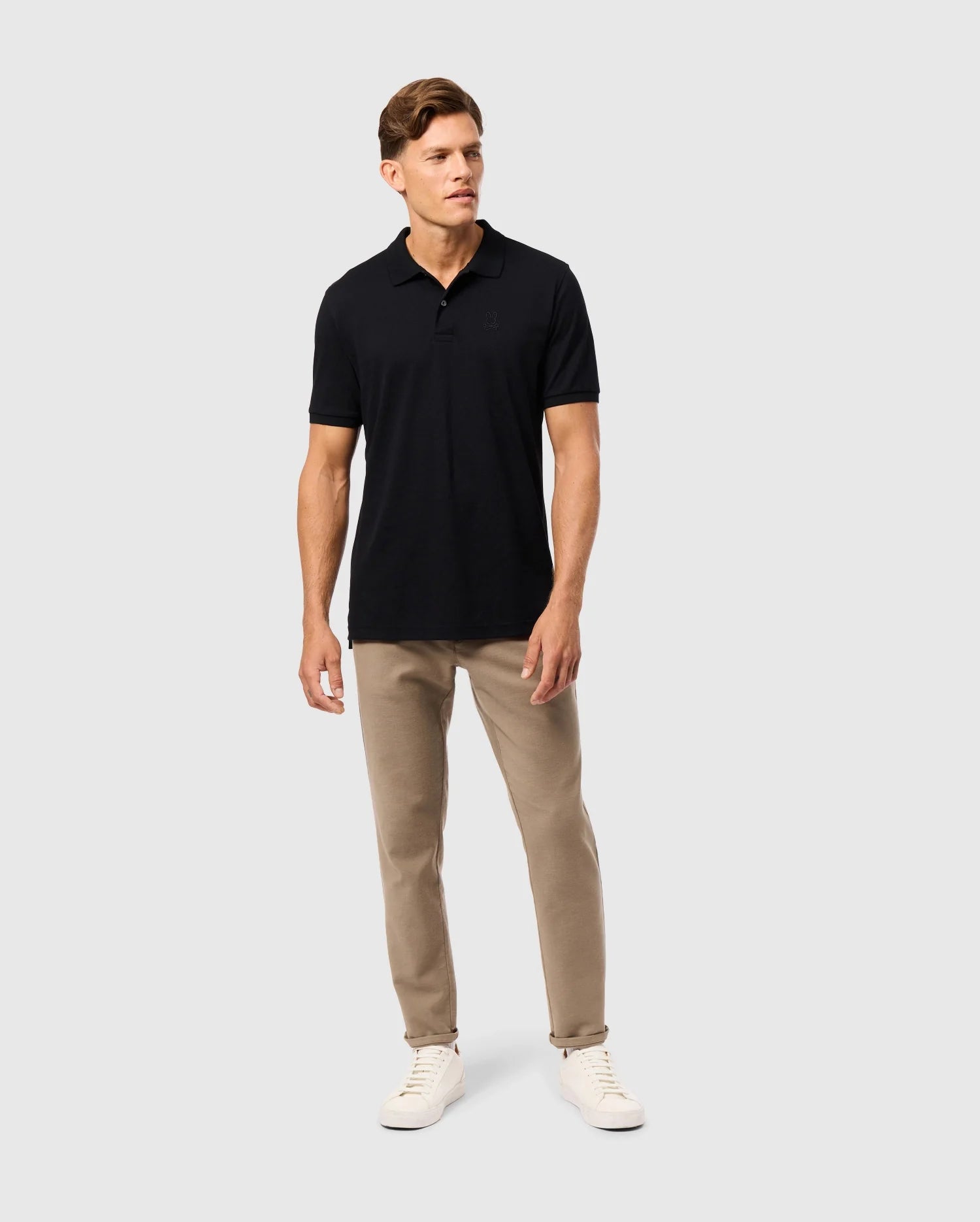 A man stands against a plain background, wearing the Psycho Bunny MENS JAMES PREMIUM PIQUE POLO SHIRT - B6K945D200 in black, paired with beige pants and white sneakers. His short hair is neatly styled as he gazes slightly to the side.