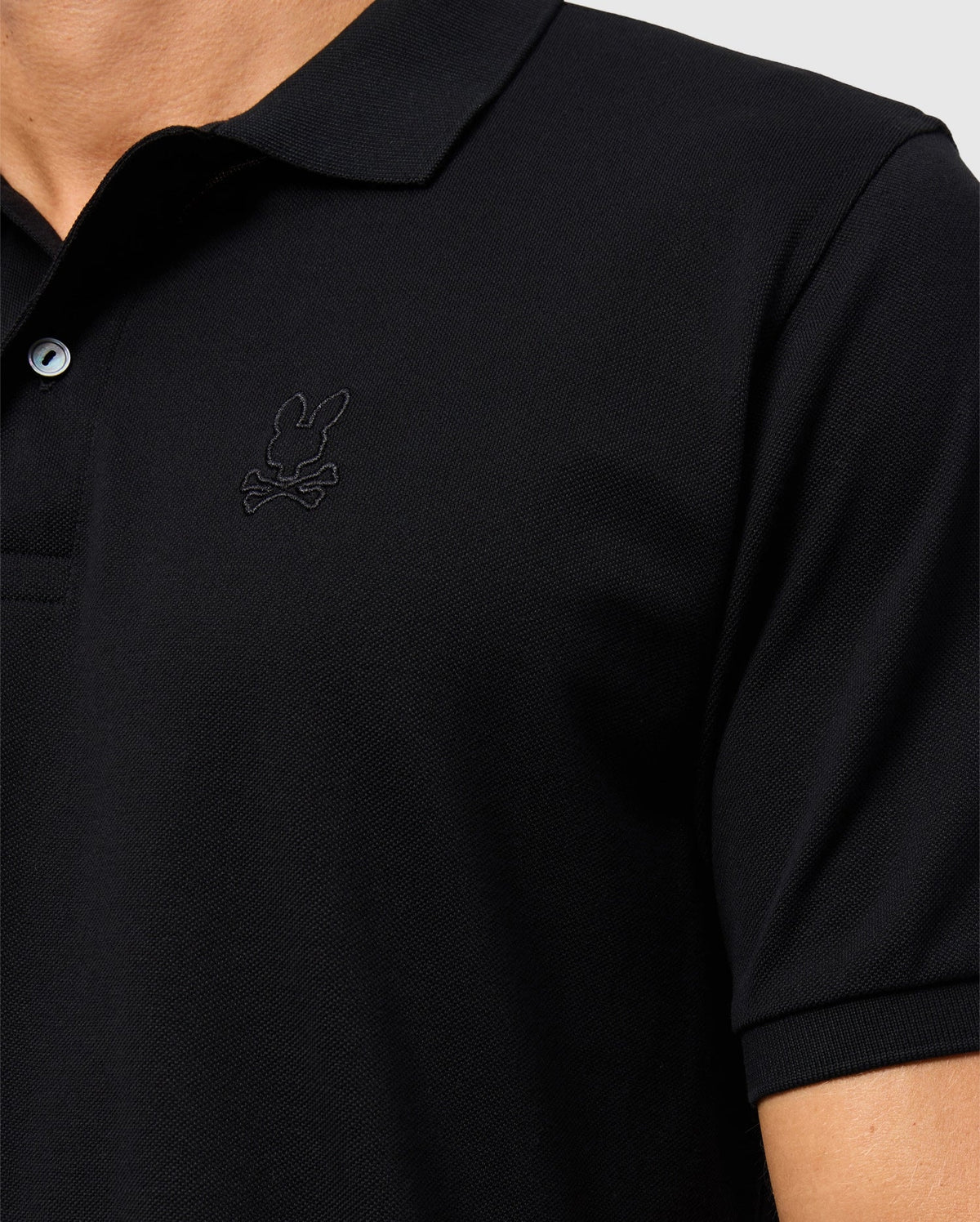 Close-up of a person wearing the Psycho Bunny MENS JAMES PREMIUM PIQUE POLO SHIRT - B6K945D200, made from premium Pima cotton. It features a rabbit and crossbones logo on the chest and includes a collar, combining style and durability against the light gray background.