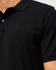 Close-up of a person wearing the Psycho Bunny MENS JAMES PREMIUM PIQUE POLO SHIRT - B6K945D200, made from premium Pima cotton. It features a rabbit and crossbones logo on the chest and includes a collar, combining style and durability against the light gray background.