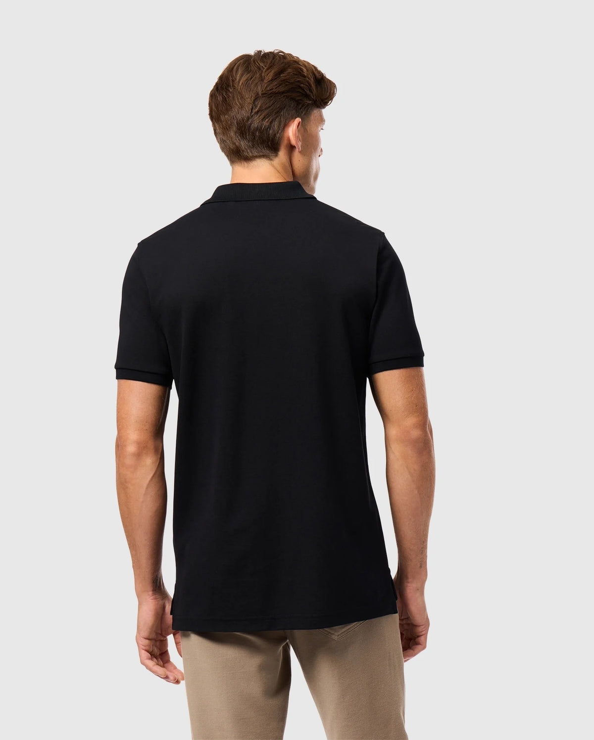 A man with brown hair is standing with his back to the camera, wearing a Psycho Bunny MENS JAMES PREMIUM PIQUE POLO SHIRT in black and beige pants against a plain light gray background.
