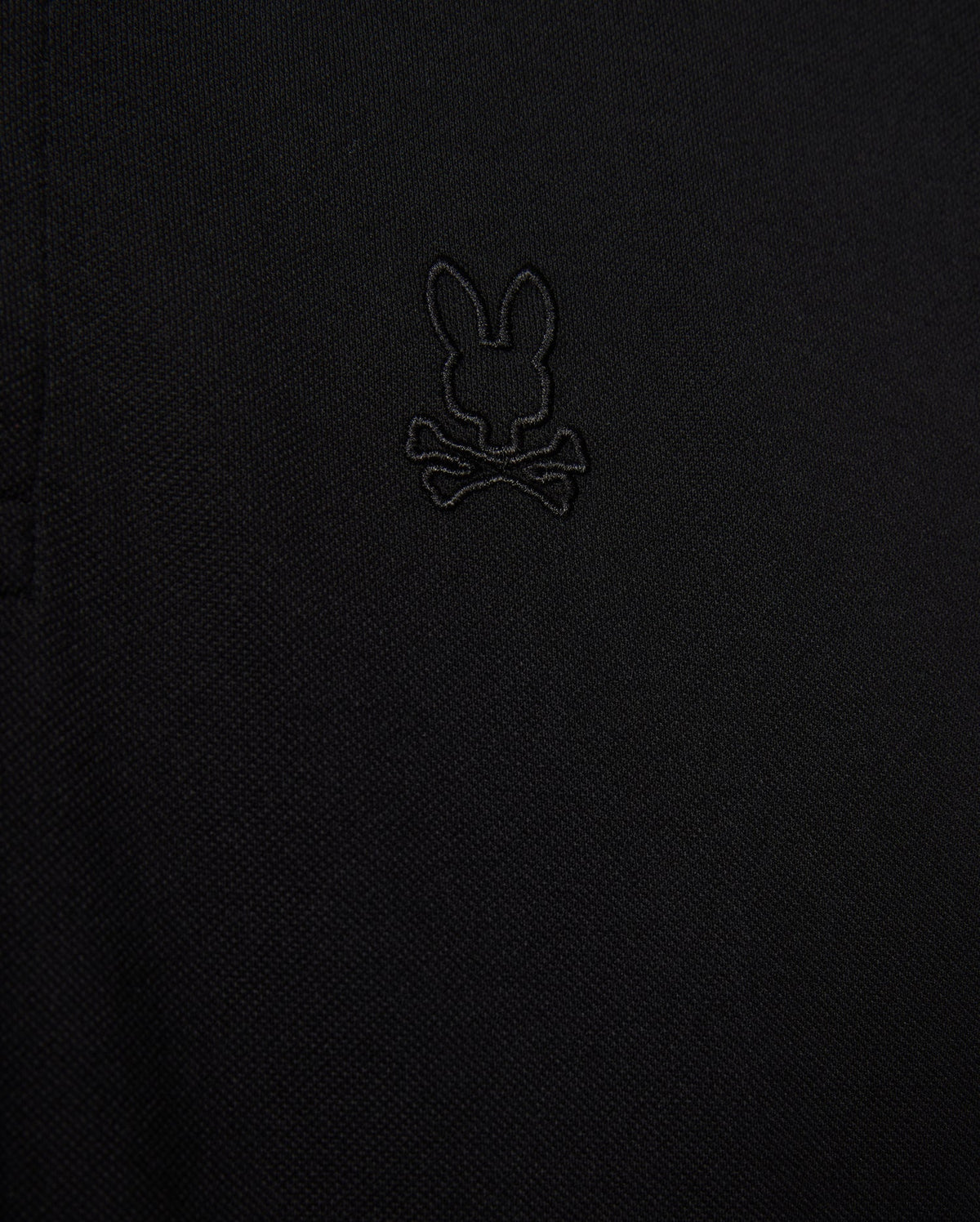 Close-up of the Psycho Bunny MENS JAMES PREMIUM PIQUE POLO SHIRT, crafted from durable black textured Pima cotton fabric and showcasing an embroidered design of a bunny head above crossed bones.