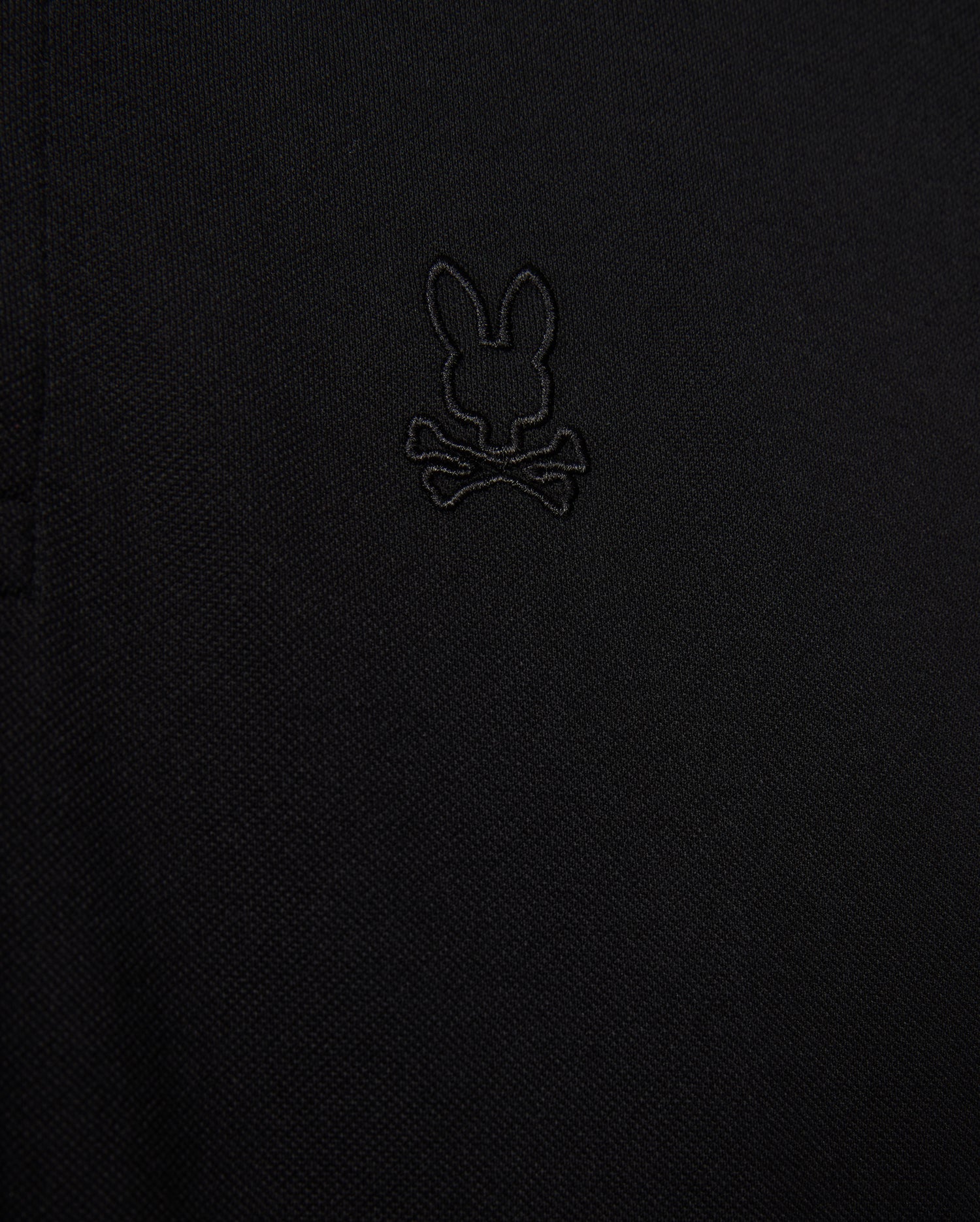 Close-up of the Psycho Bunny MENS JAMES PREMIUM PIQUE POLO SHIRT, crafted from durable black textured Pima cotton fabric and showcasing an embroidered design of a bunny head above crossed bones.