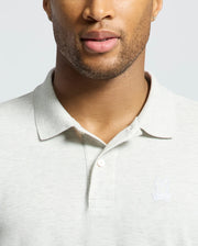 A man wears a Psycho Bunny MENS JAMES PIQUE POLO - B6K945D200 in light gray with an embroidered rabbit and crossed bones logo. His partially visible face shows trimmed facial hair. Made from soft Pima cotton, this shirt offers a durable, slim fit.