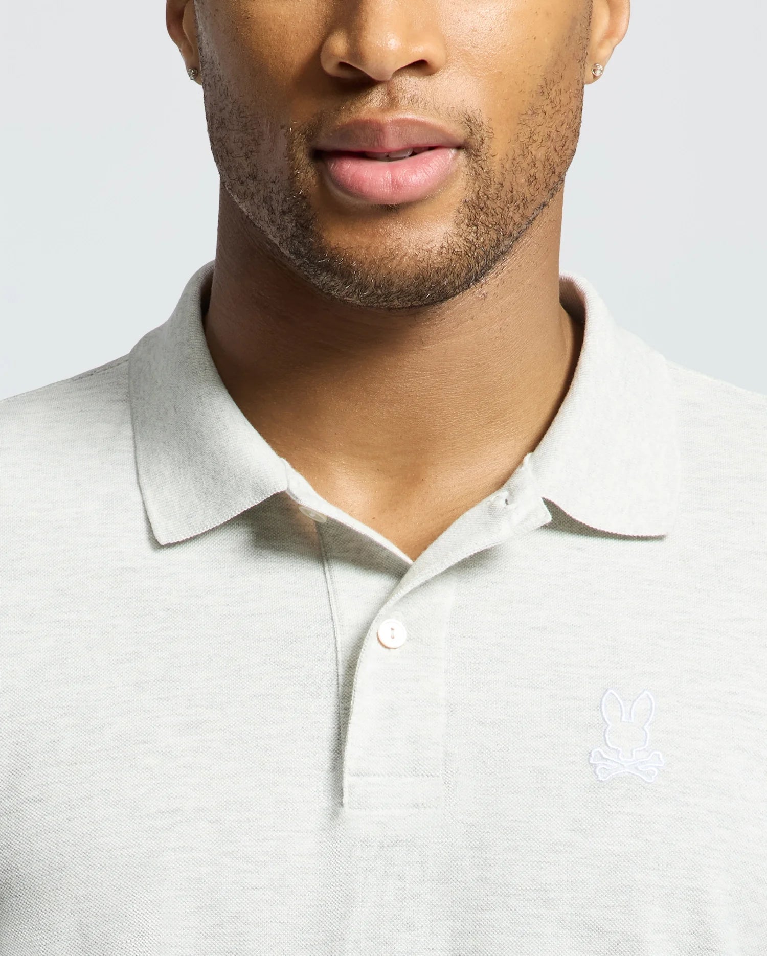 A man wears a Psycho Bunny MENS JAMES PIQUE POLO - B6K945D200 in light gray with an embroidered rabbit and crossed bones logo. His partially visible face shows trimmed facial hair. Made from soft Pima cotton, this shirt offers a durable, slim fit.