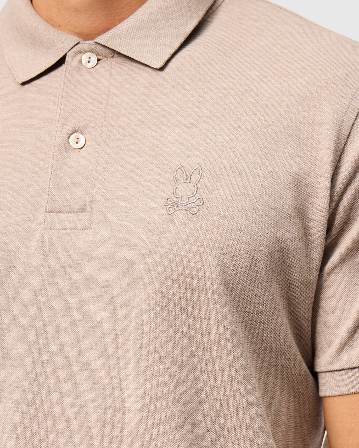 A person is wearing the MENS JAMES PREMIUM PIQUE POLO SHIRT - B6K945D200 by Psycho Bunny, made from soft Pima cotton. This beige polo features a bunny and crossbones logo embroidered on the left chest, with a collar and two buttons for style and durability.