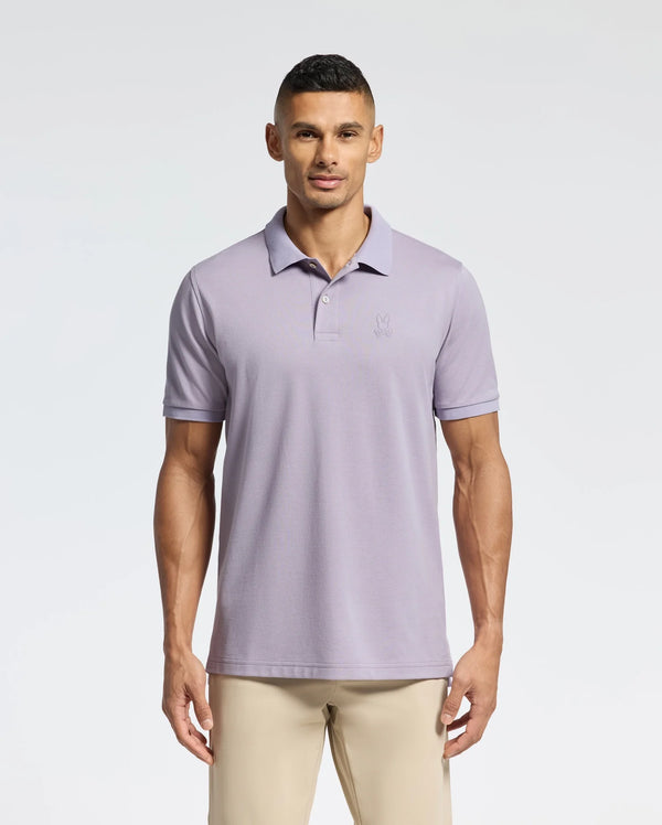 A person stands against a plain white background wearing the Psycho Bunny MENS JAMES PIQUE POLO - B6K945D200 in light purple, paired with beige pants. The durable Pima cotton shirt has a subtle logo on the chest and a crease-resistant collar.