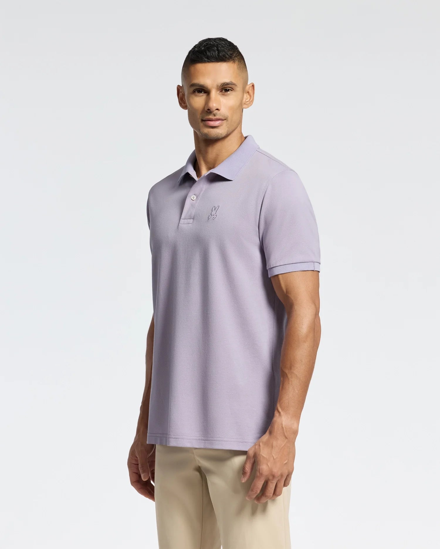 A person in a Psycho Bunny Slim Fit MENS JAMES PIQUE POLO - B6K945D200 in light purple and beige pants stands against a plain white background. Made from durable Pima cotton, they gaze at the camera with a neutral expression.