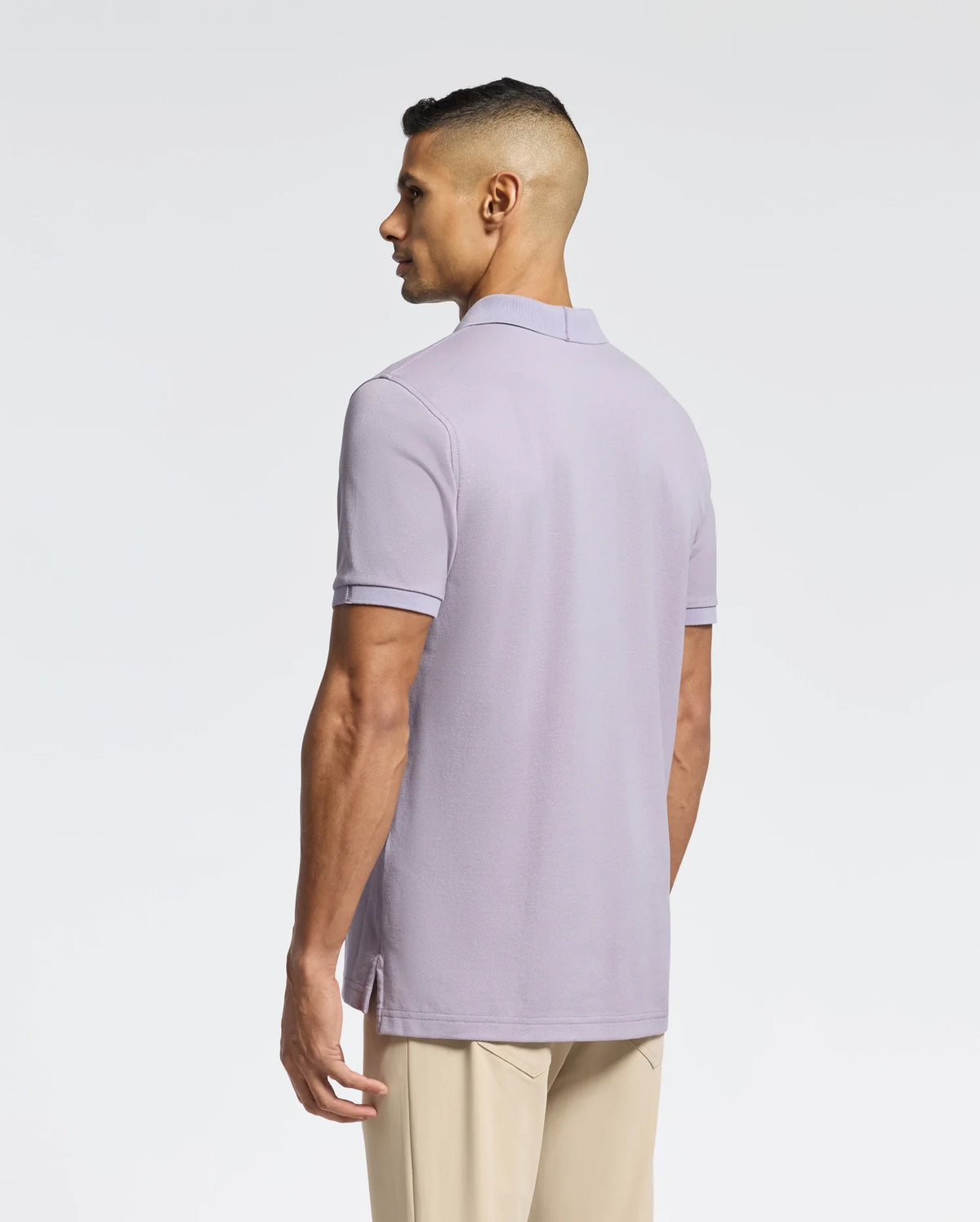 A man with short dark hair, back to the camera, wears a Psycho Bunny MENS JAMES PIQUE POLO - B6K945D200 in light purple made from durable Pima cotton and beige pants against a plain white background.