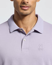 A person is wearing the Psycho Bunny MENS JAMES PIQUE POLO - B6K945D200 in light purple. It's a slim-fit polo with a bunny head and crossbones logo embroidered on the left chest. Made of durable Pima cotton, it features a collar and two buttons, with the top button undone for style.
