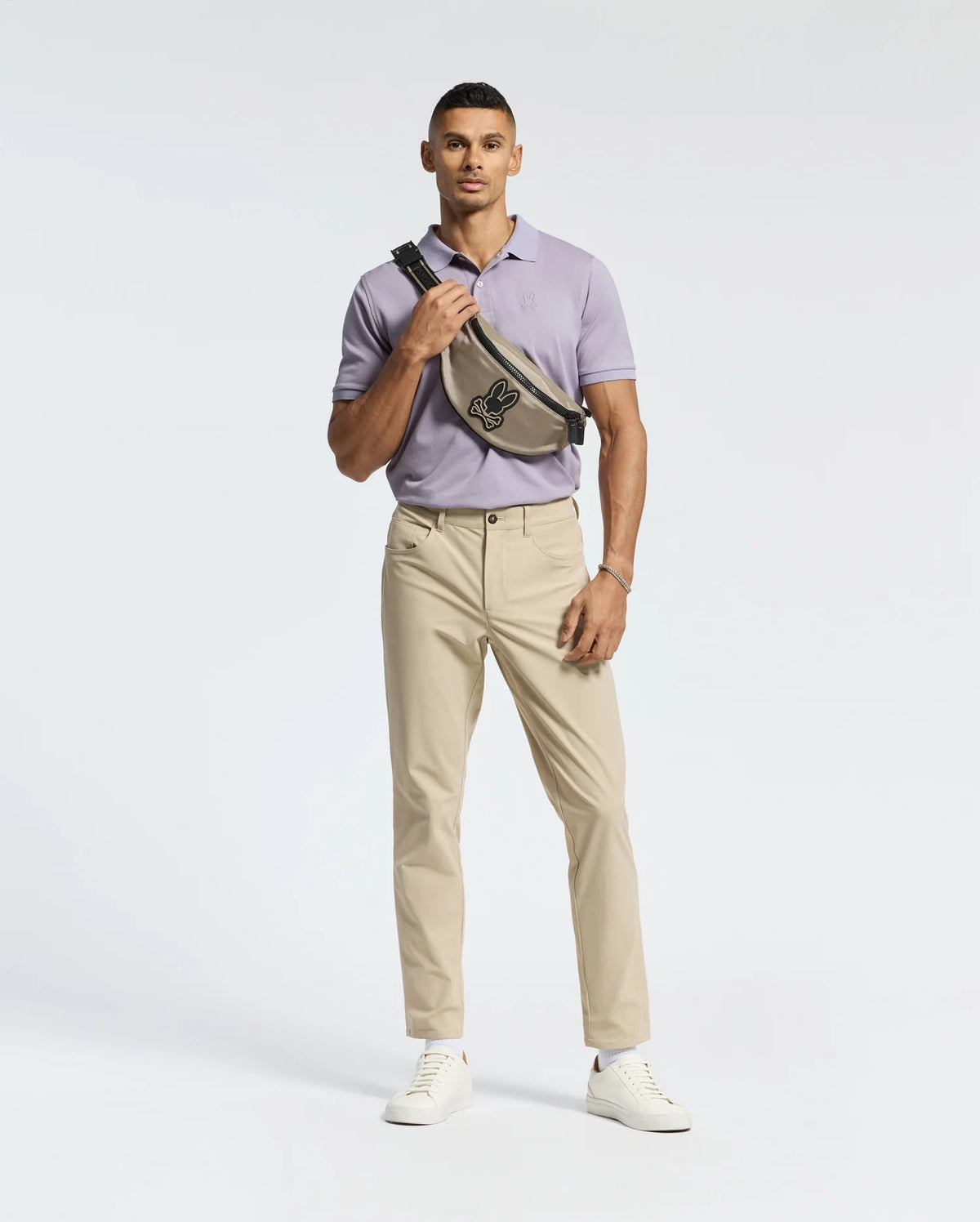 A man stands against a white background, wearing a light purple Psycho Bunny MENS JAMES PIQUE POLO - B6K945D200 and beige pants. He has a black graphic crossbody bag, white sneakers, and a wrist accessory on his right arm.