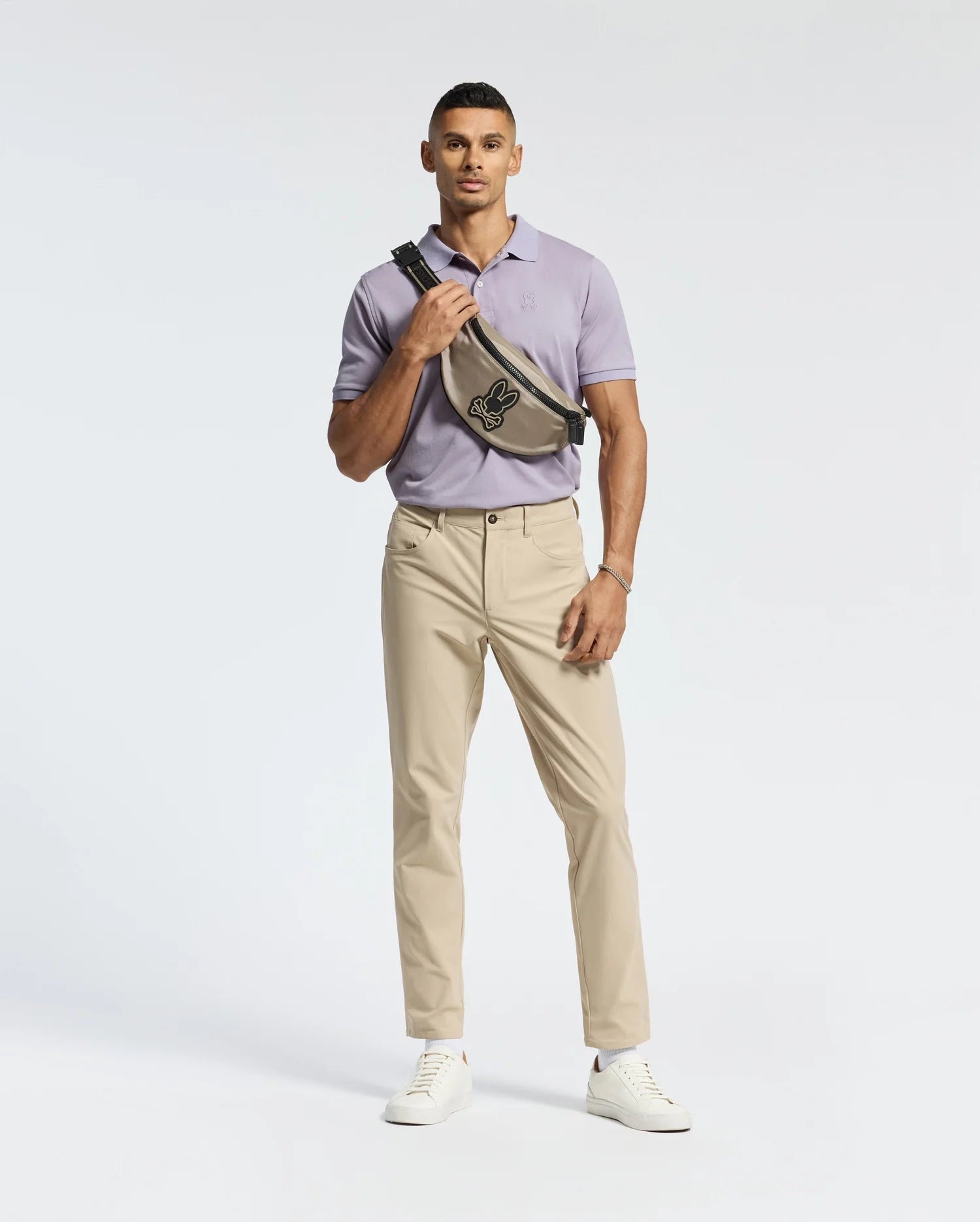 A man stands against a white background, wearing a light purple Psycho Bunny MENS JAMES PIQUE POLO - B6K945D200 and beige pants. He has a black graphic crossbody bag, white sneakers, and a wrist accessory on his right arm.