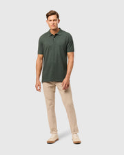 A man stands against a plain background wearing the Psycho Bunny Men's James Premium Pique Polo Shirt in dark green, paired with beige pants and light-colored sneakers. His hands rest relaxed at his sides as he gazes forward confidently.
