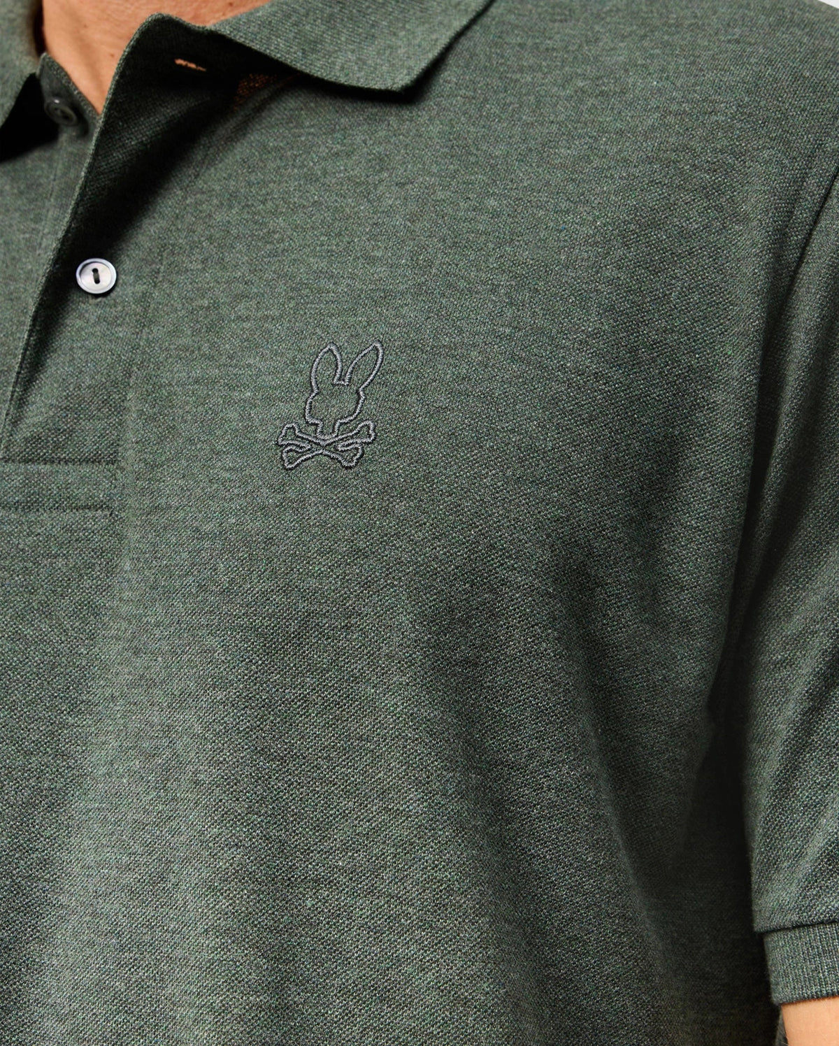 Close-up of a person wearing the Psycho Bunny Mens James Premium Pique Polo Shirt - B6K945D200 in dark green, showcasing an embroidered bunny with crossbones logo on the chest. This high-quality polo features a buttoned collar and is crafted from textured fabric, offering both style and comfort.