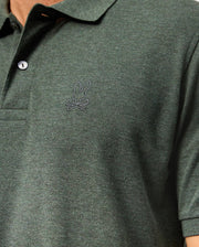 Close-up of a person wearing the Psycho Bunny Mens James Premium Pique Polo Shirt - B6K945D200 in dark green, showcasing an embroidered bunny with crossbones logo on the chest. This high-quality polo features a buttoned collar and is crafted from textured fabric, offering both style and comfort.