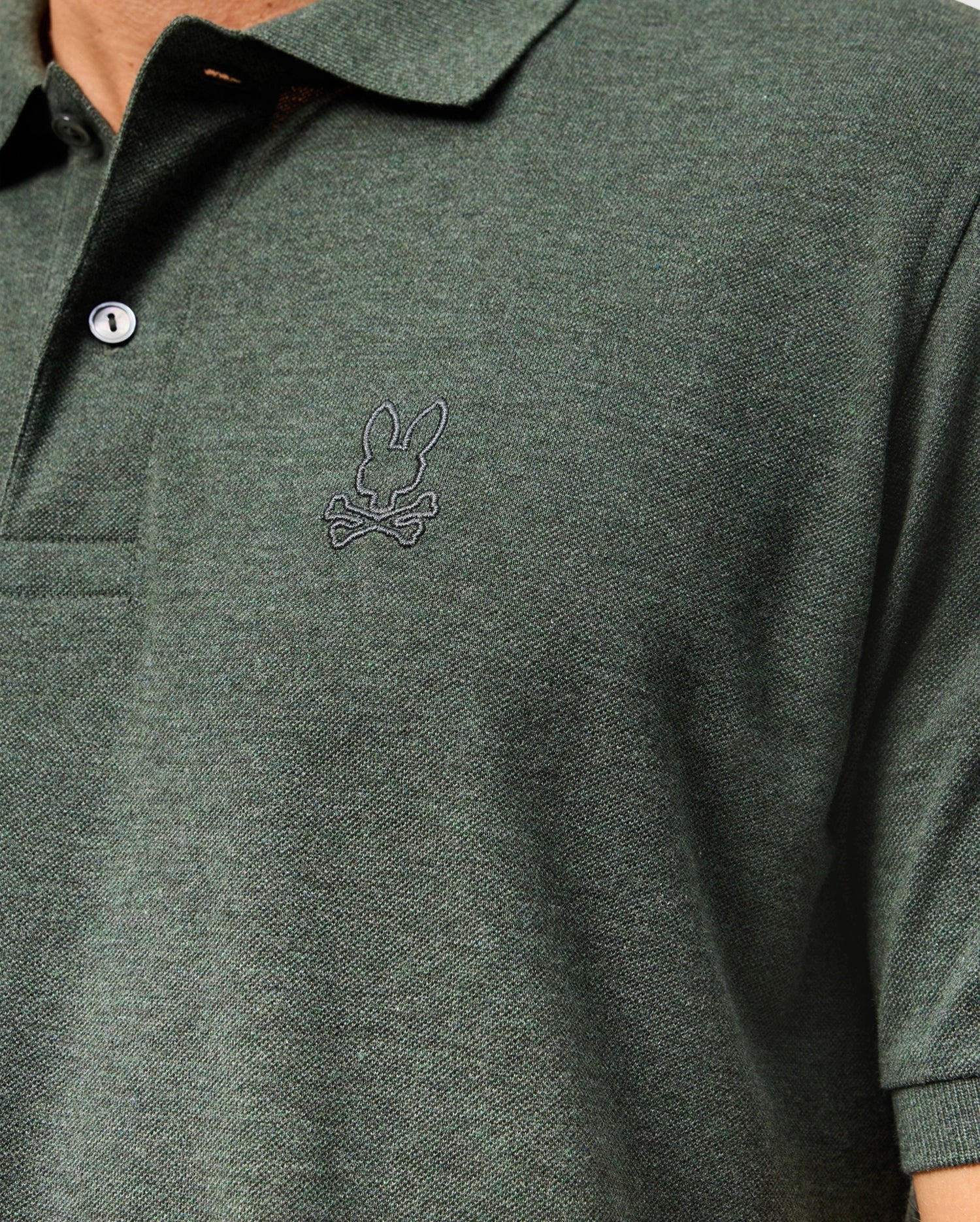 Close-up of a person wearing the Psycho Bunny Mens James Premium Pique Polo Shirt - B6K945D200 in dark green, showcasing an embroidered bunny with crossbones logo on the chest. This high-quality polo features a buttoned collar and is crafted from textured fabric, offering both style and comfort.