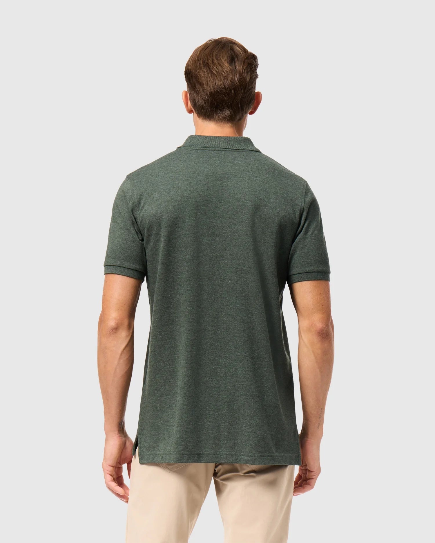A man in a Psycho Bunny James Premium Pique Polo Shirt, made from lightweight pima cotton, and beige pants stands with his back to the camera against a plain gray background.