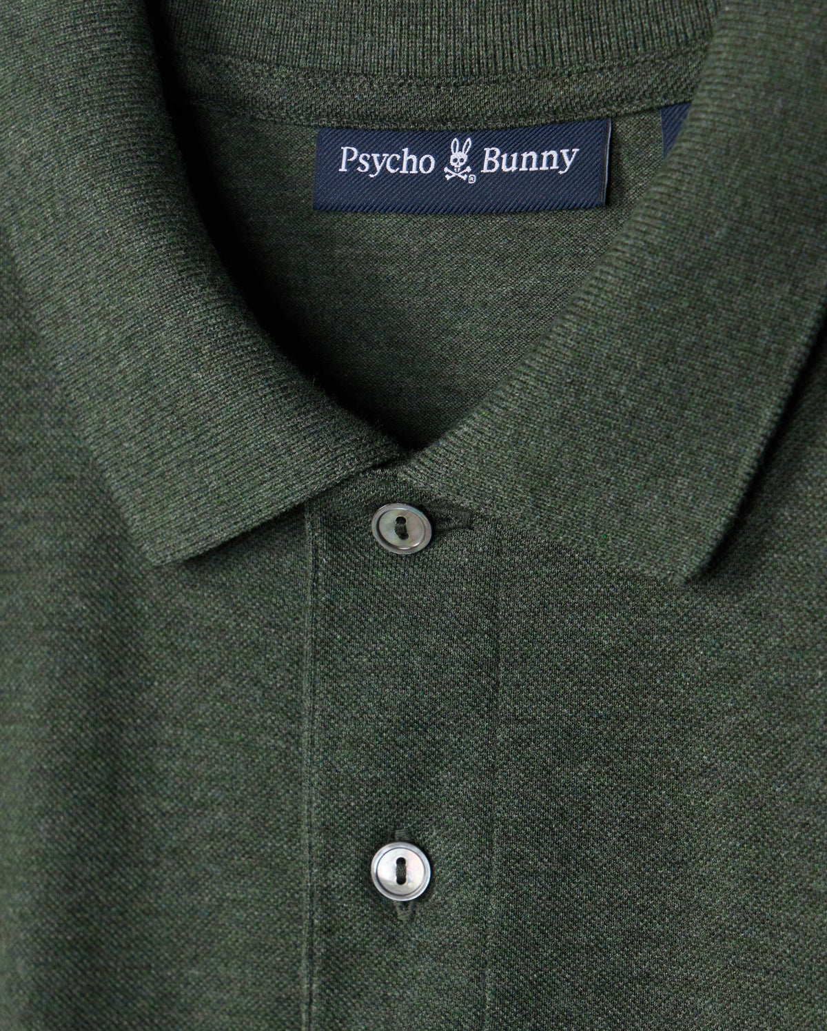 Close-up of a dark green premium pique polo shirt featuring a collar and two buttons. The label displays the brand "Psycho Bunny" in white letters on a dark blue tag, showcasing the high-quality craftsmanship of the Men's James Premium Pique Polo Shirt made from lightweight pima cotton.